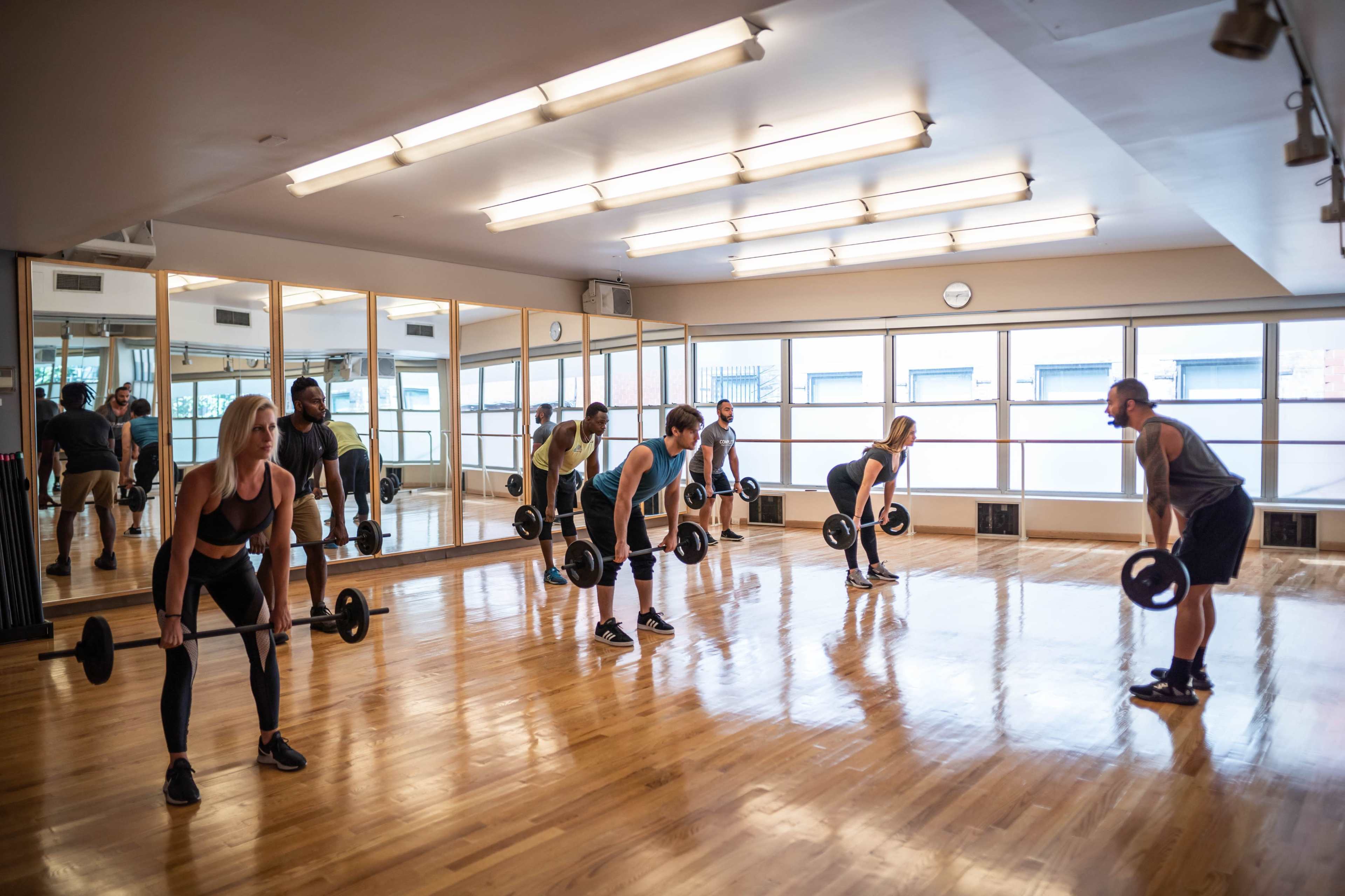 A group of individuals is performing deadlifts with barbell weights in a brightly lit gym studio with mirrors on one wall.