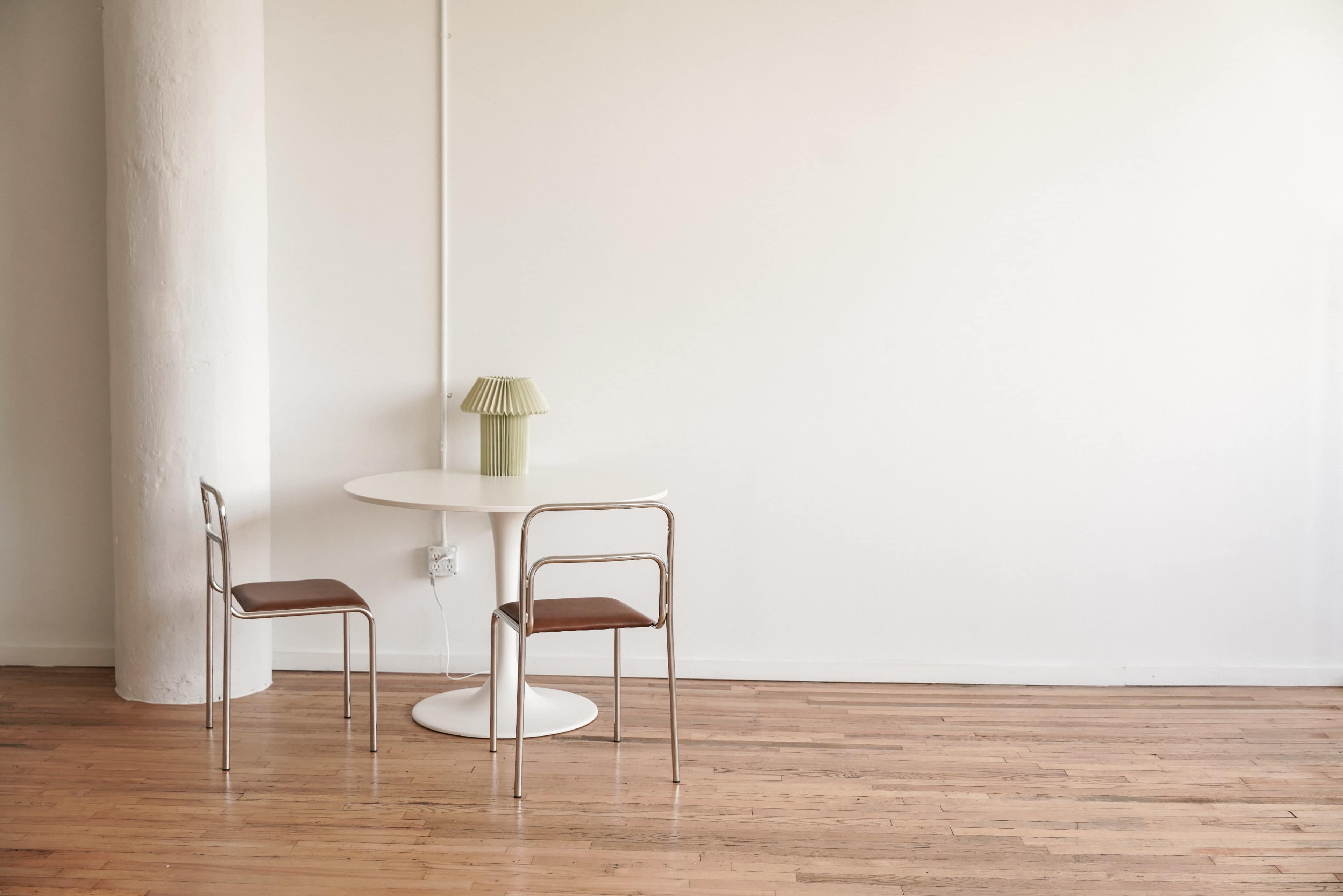 A round, white table with two chairs and a green lamp stands in a minimalist room with wooden flooring.