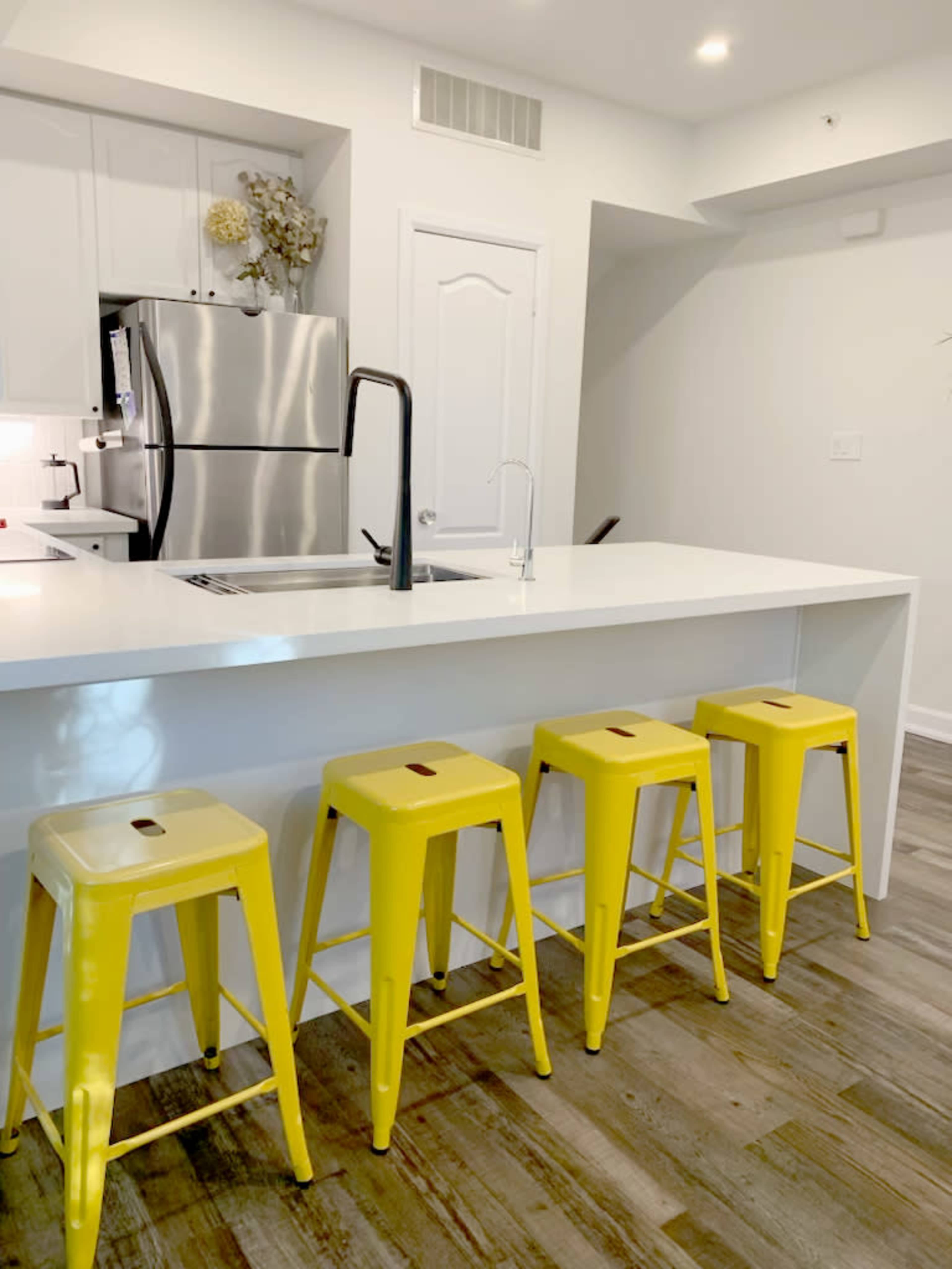 The image shows a modern kitchen with a white countertop and four yellow metal stools arranged in front of it.