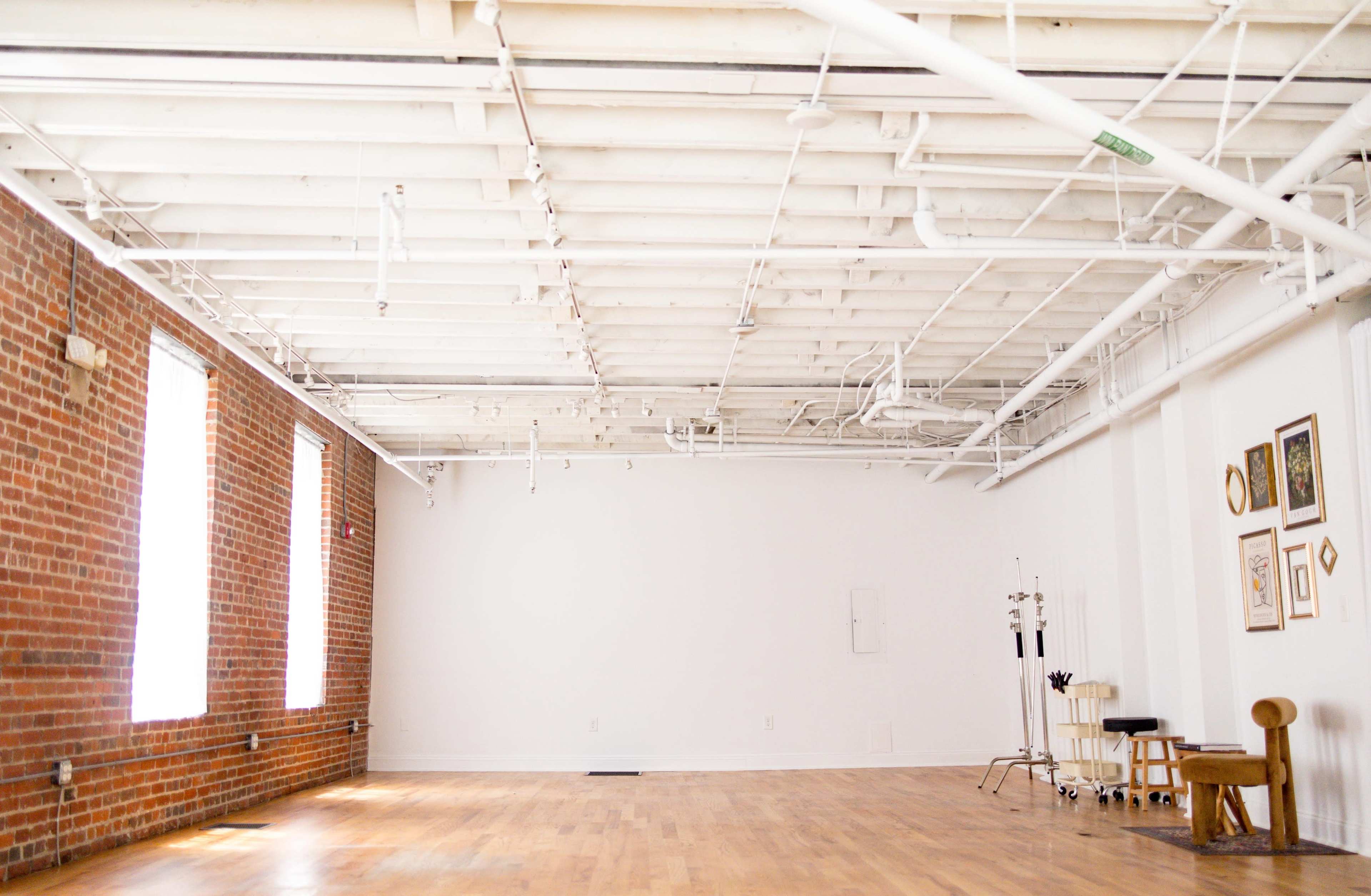A spacious, empty room with exposed brick walls, wooden flooring, and several windows allowing natural light to enter.
