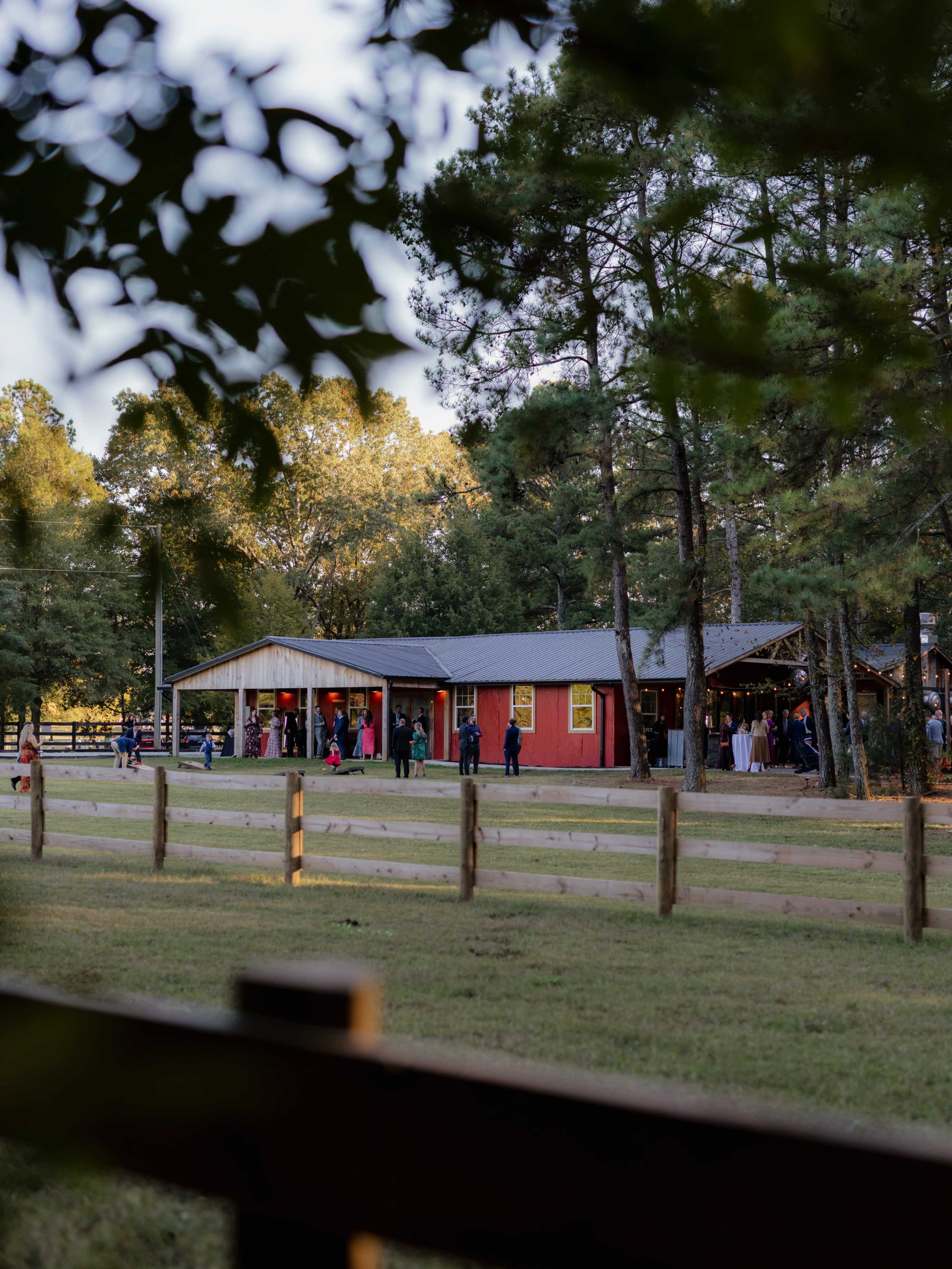 A red barn-style building is surrounded by trees and a wooden fence, with people gathered outside.