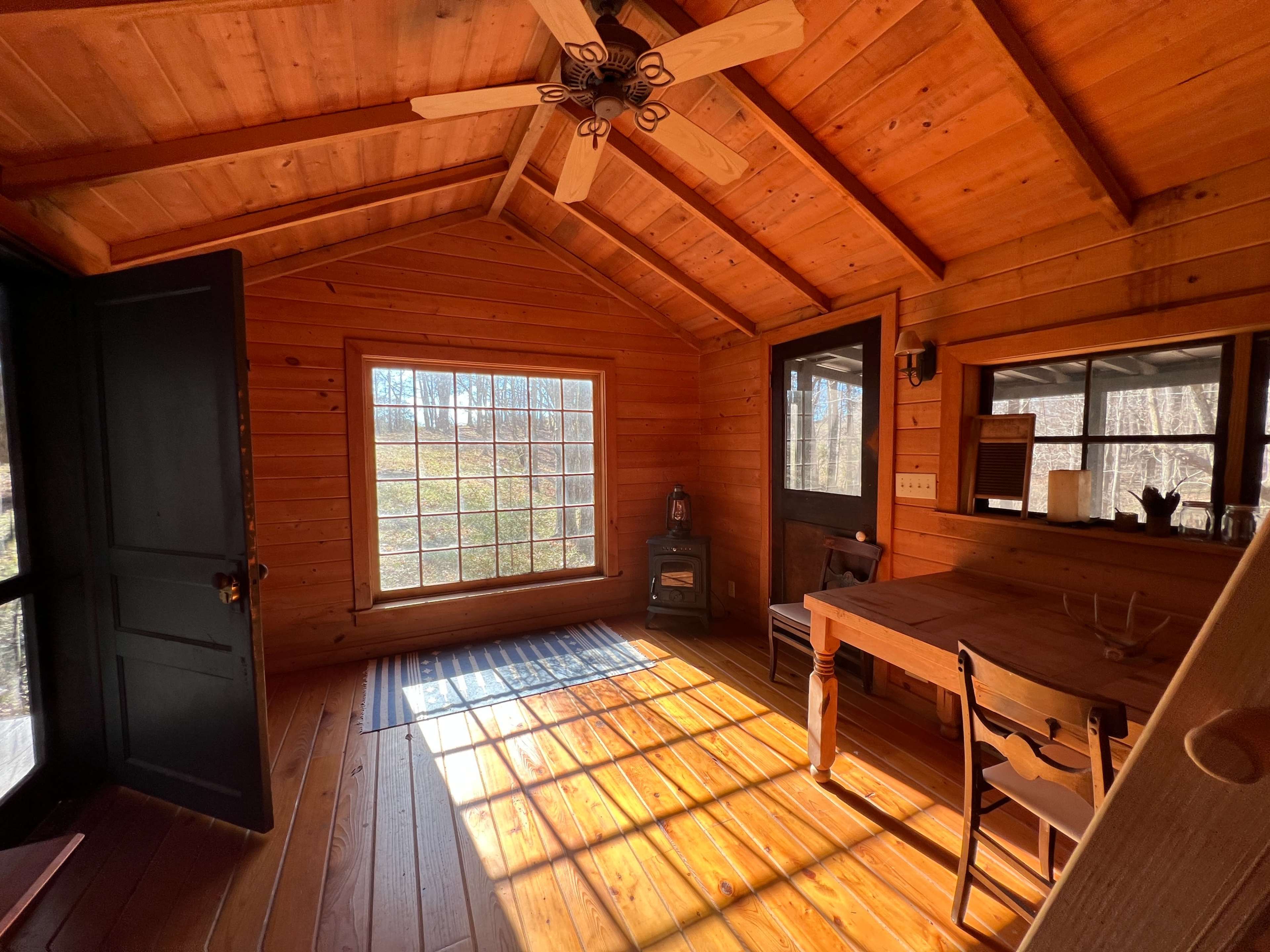 The room features wooden walls and a ceiling with exposed beams, a large window allowing natural light, a wooden table, and a small stove.