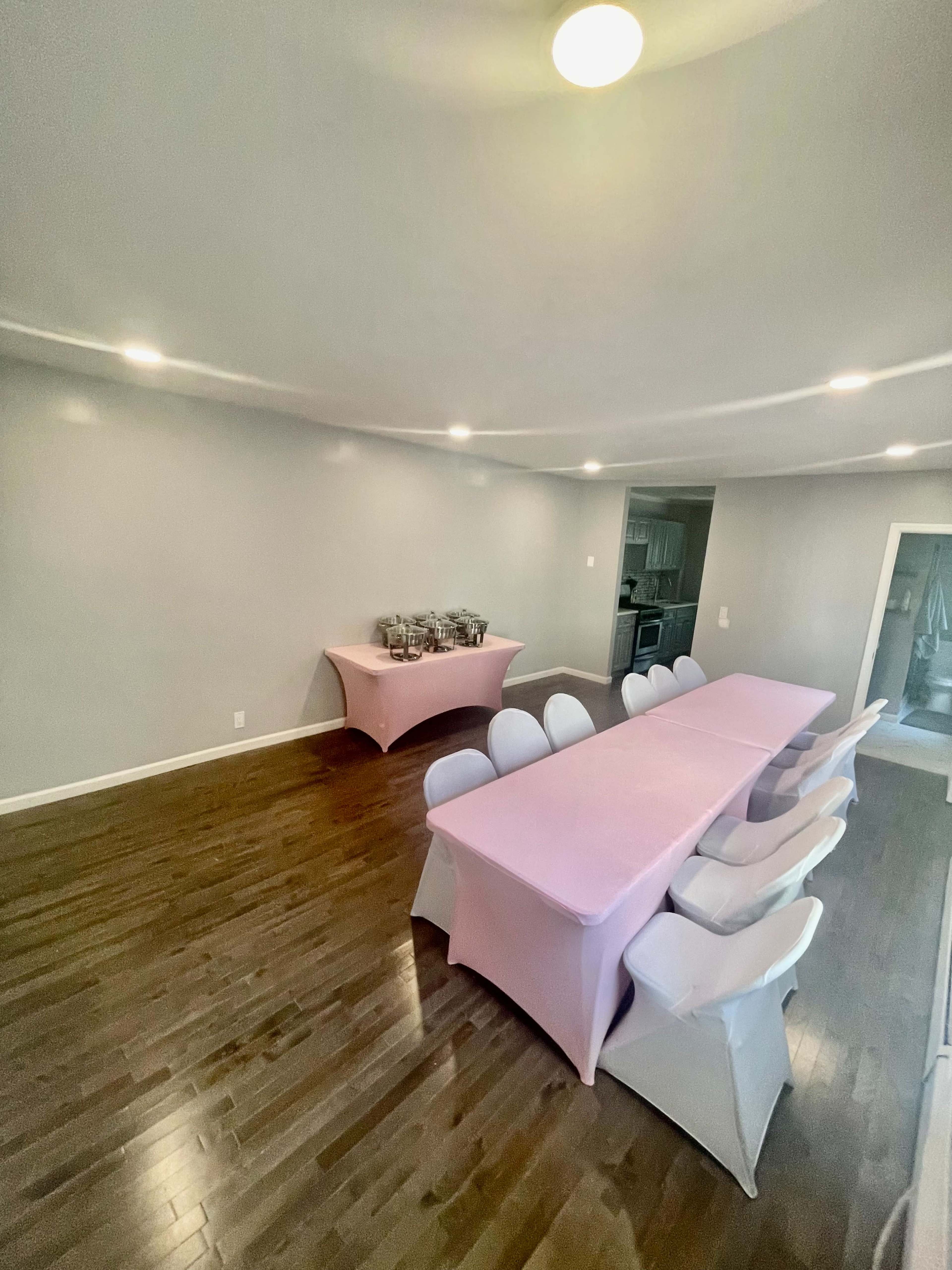 A spacious dining area features a long table covered with a pink tablecloth and surrounded by white chairs, with additional tables in the background.