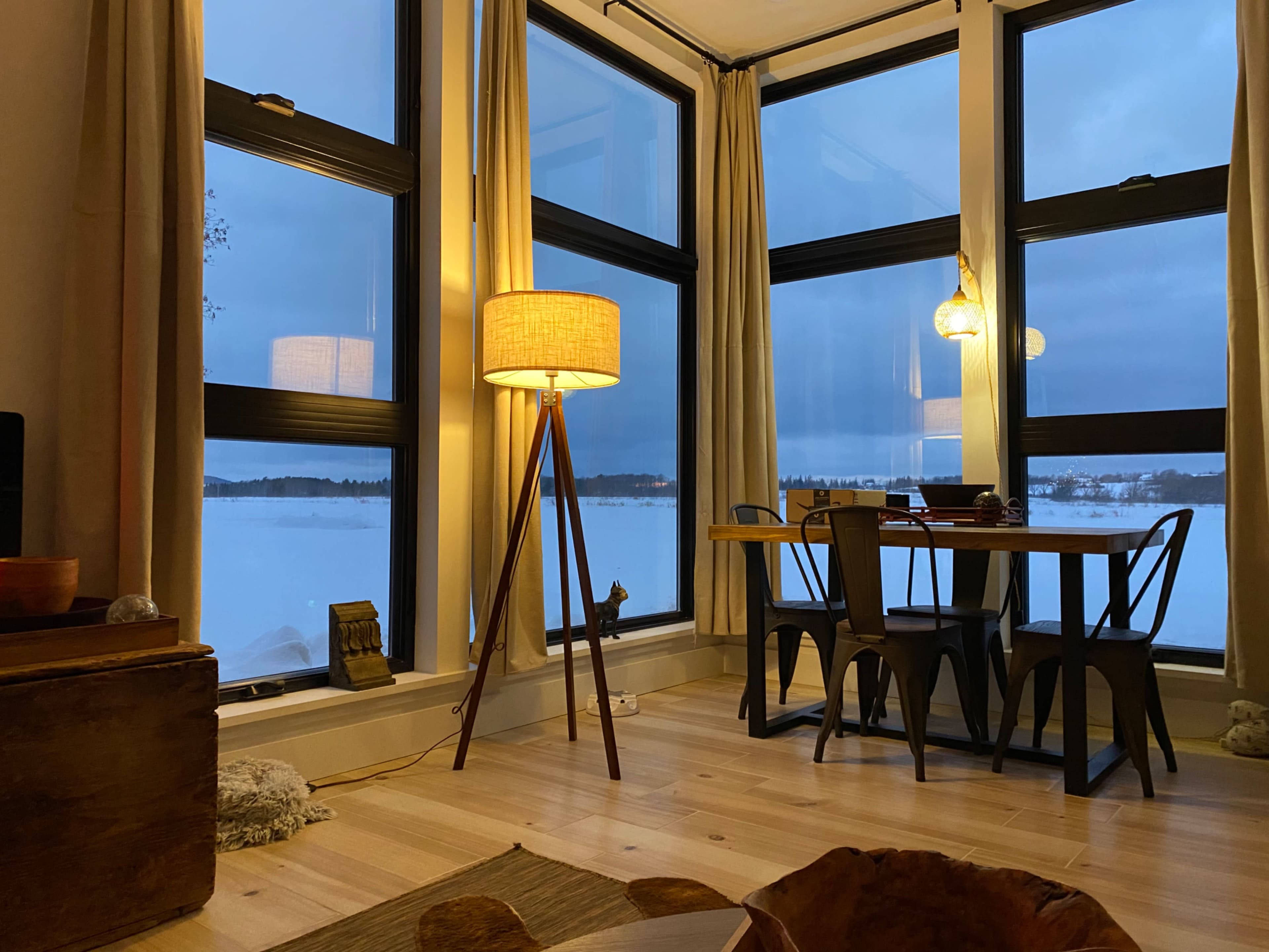 A cozy dining area with a wooden table and black chairs, surrounded by large windows that offer a view of a snowy landscape outside.