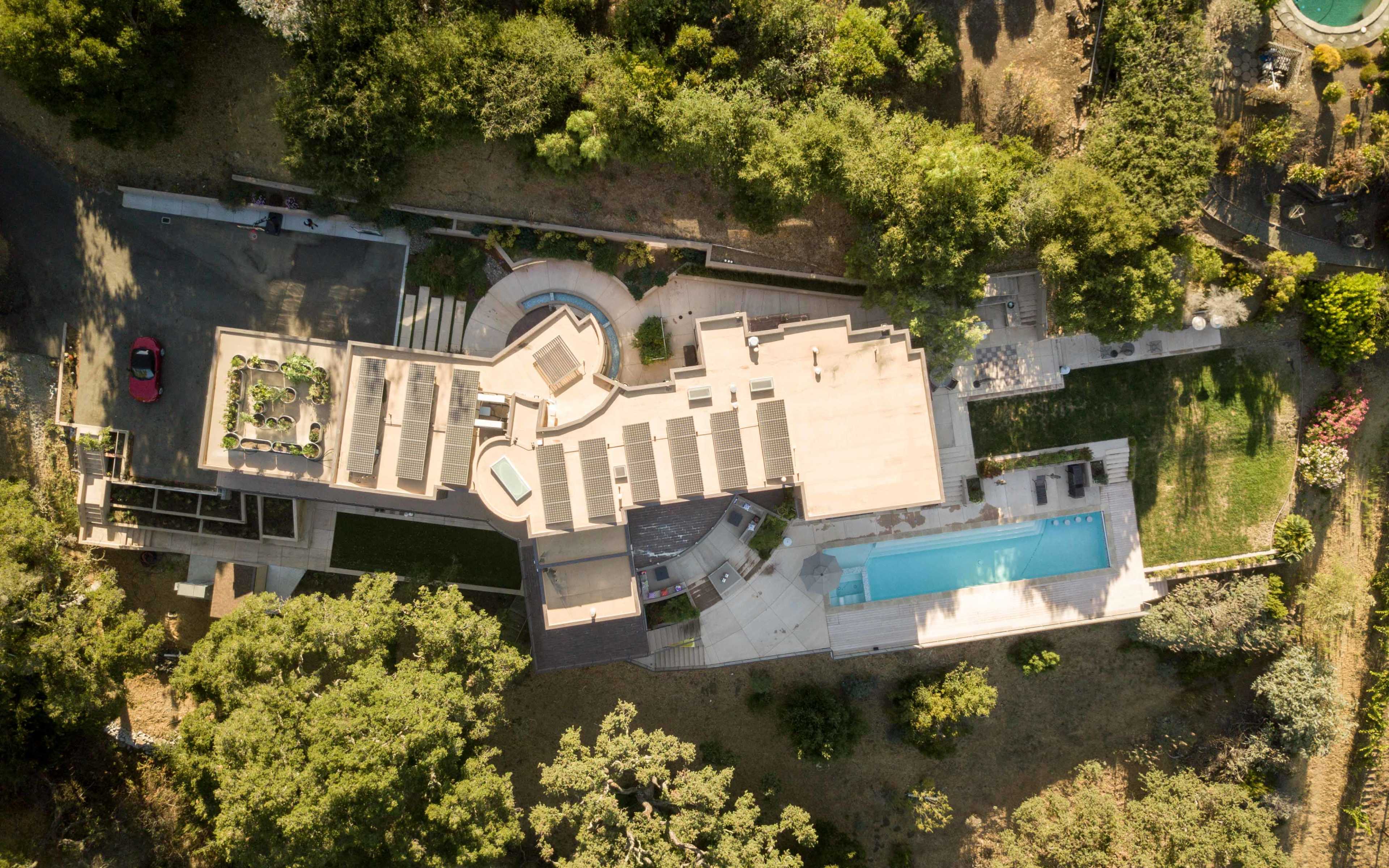 An aerial view showcases a modern house with a swimming pool, outdoor lounge areas, and landscaped gardens surrounded by trees.