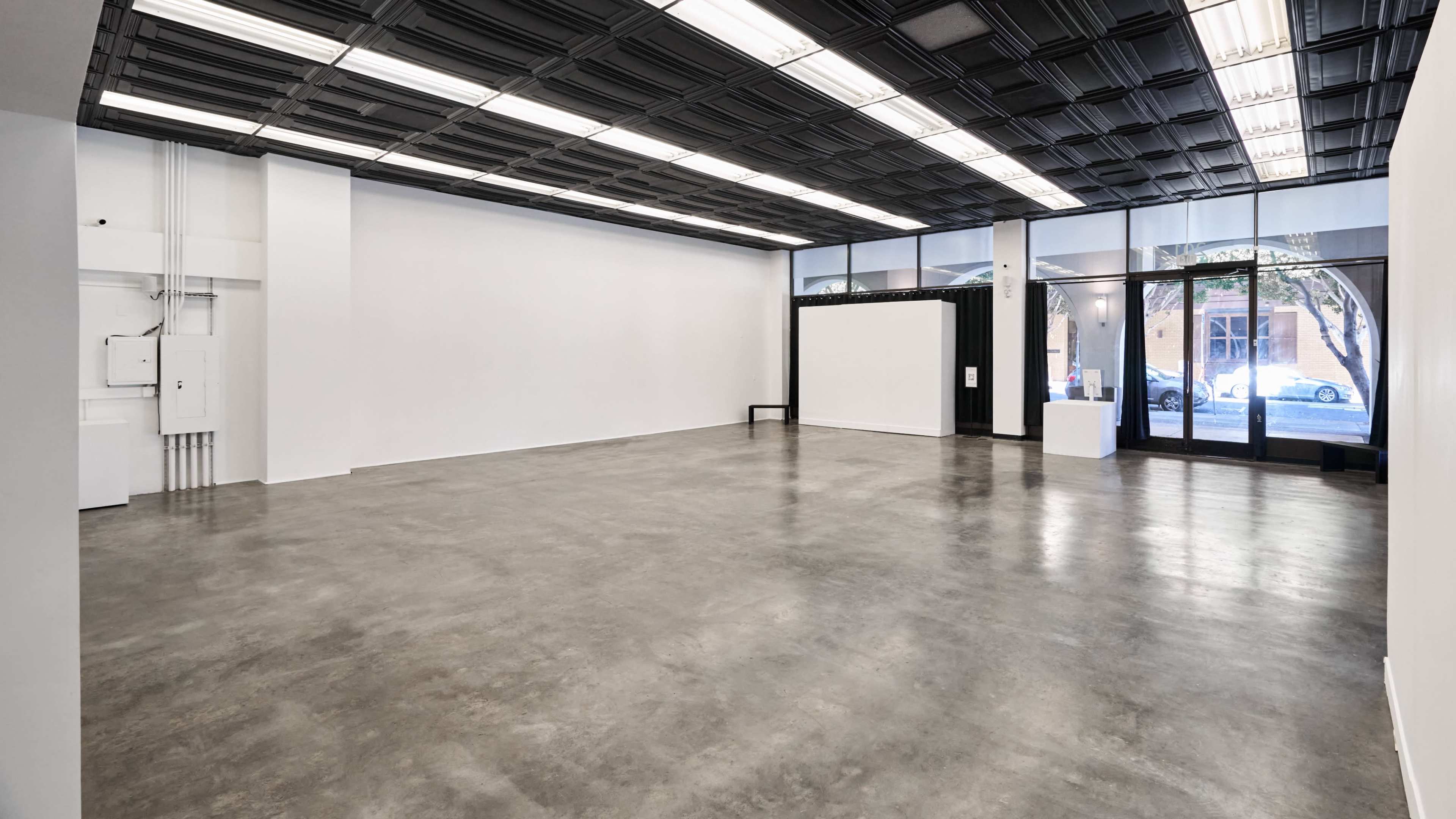 The image shows a spacious, empty art gallery with polished concrete floors and large windows allowing natural light to enter.