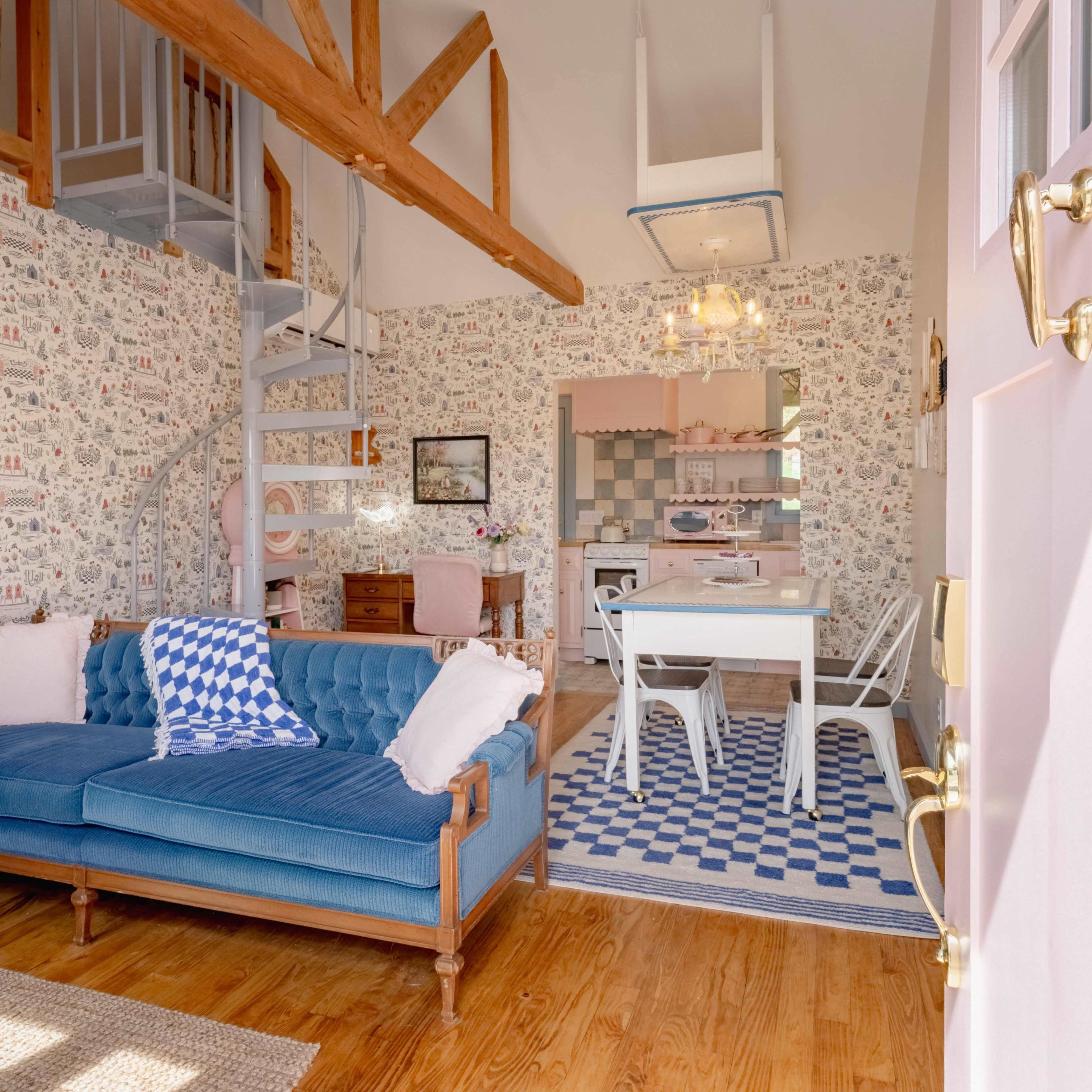 The image depicts a cozy room featuring a blue sofa, a spiral staircase, a dining area with a white table and chairs, and patterned wallpaper.