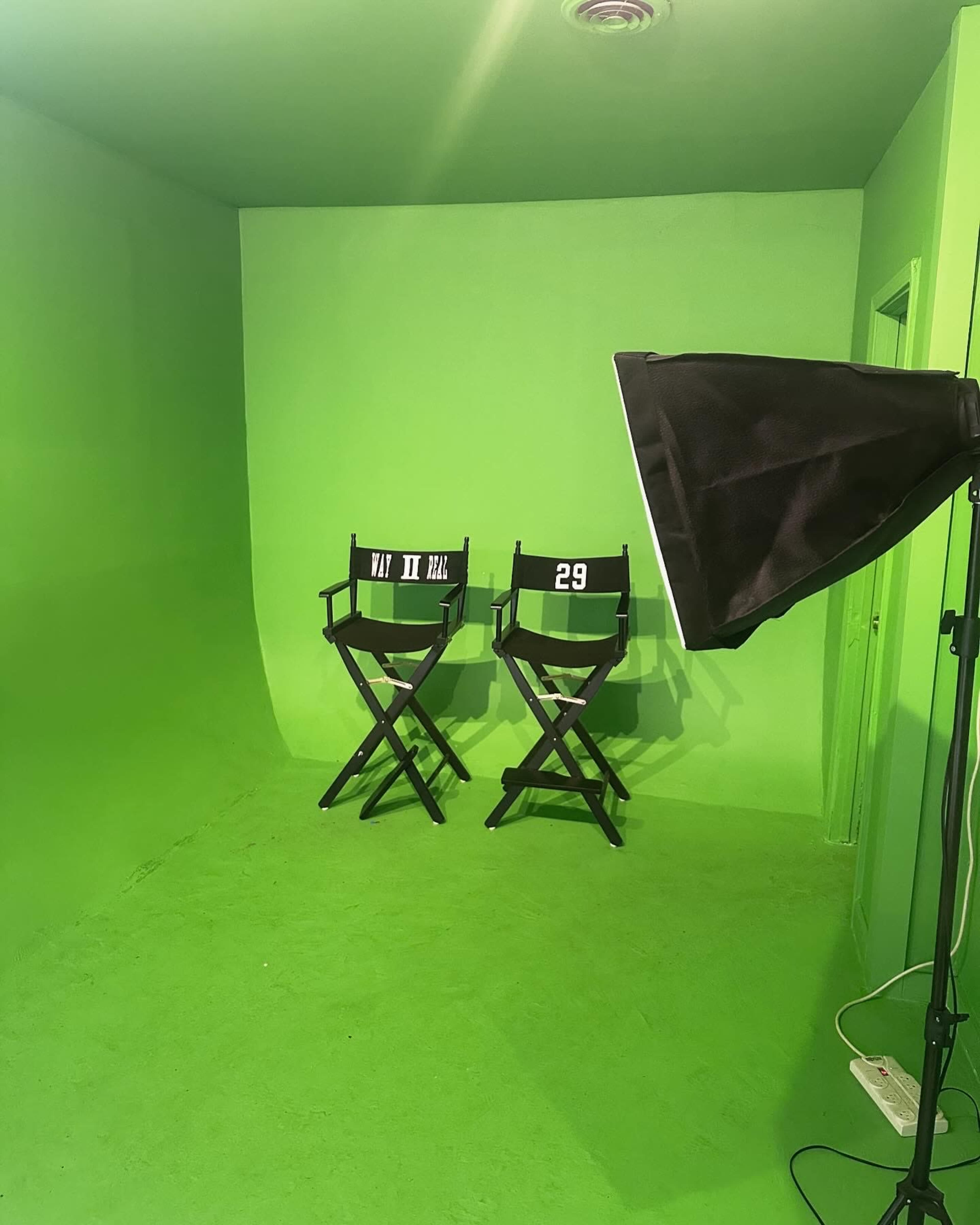 The image shows a green screen room with two director's chairs and a light source in the corner.