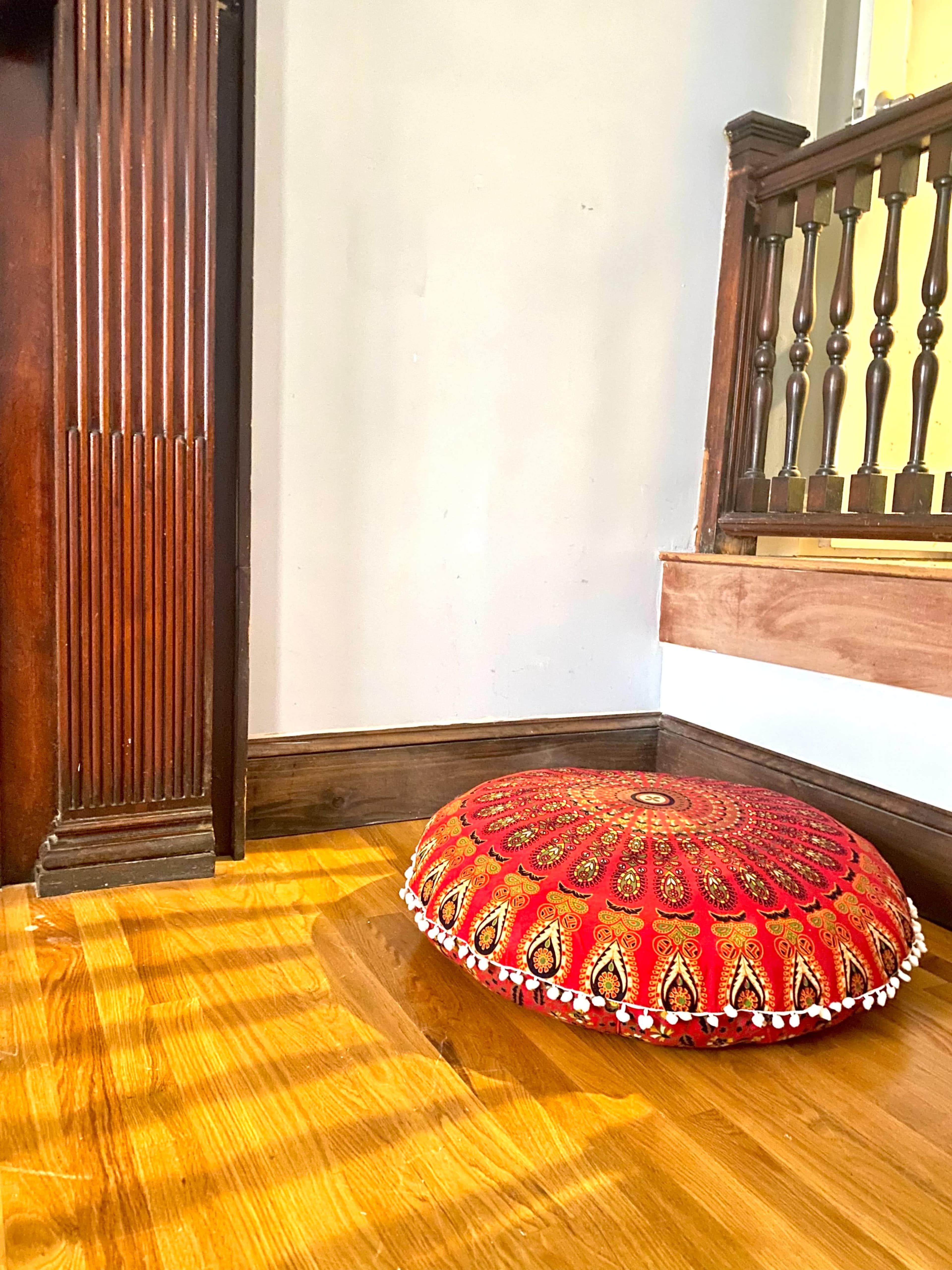 A round, decorative cushion with a red fabric and intricate patterns sits on a wooden floor in a corner near a staircase.