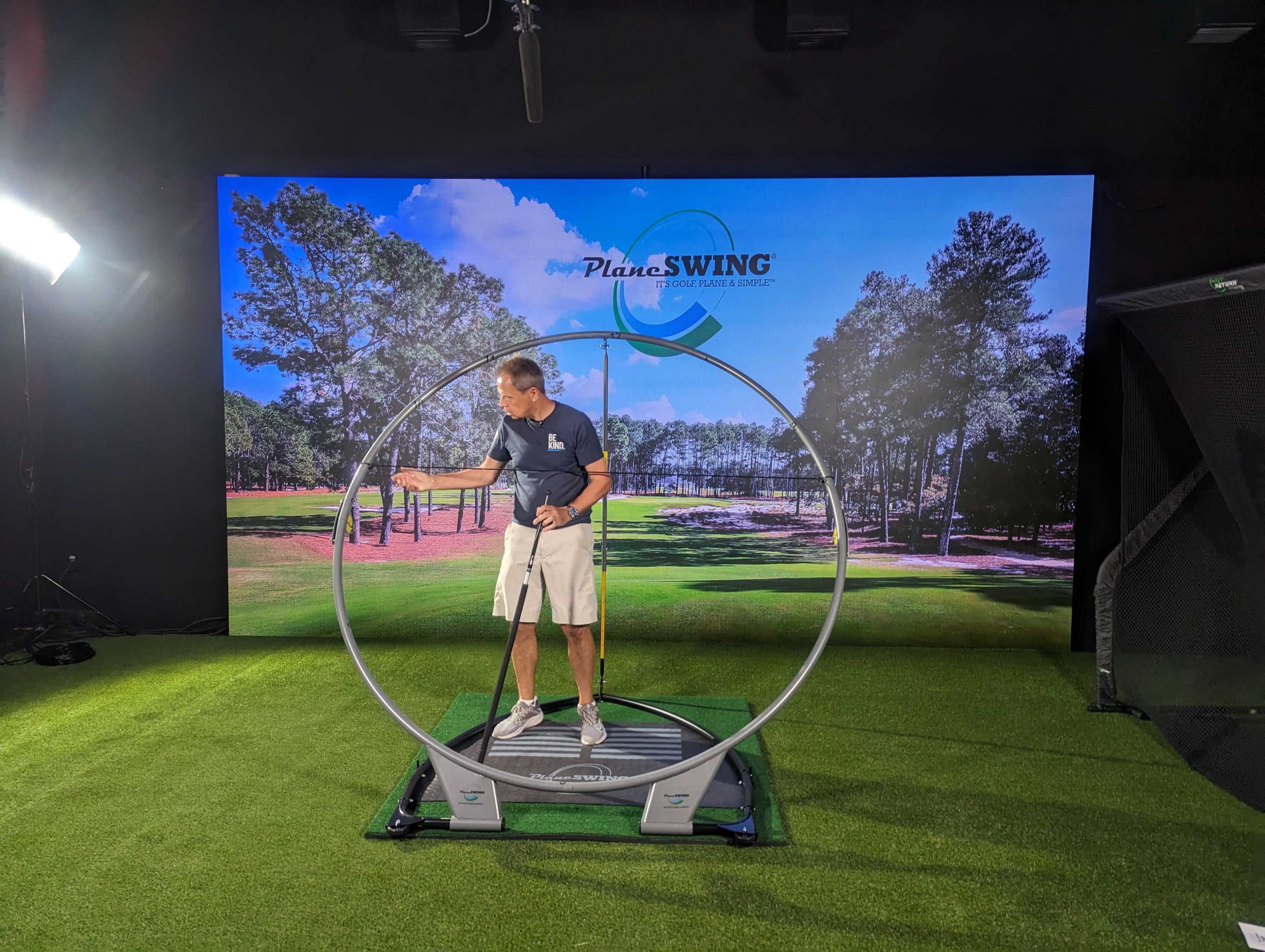 A golfer is demonstrating a swing technique using a training device in a golf simulation area with a backdrop of a golf course.