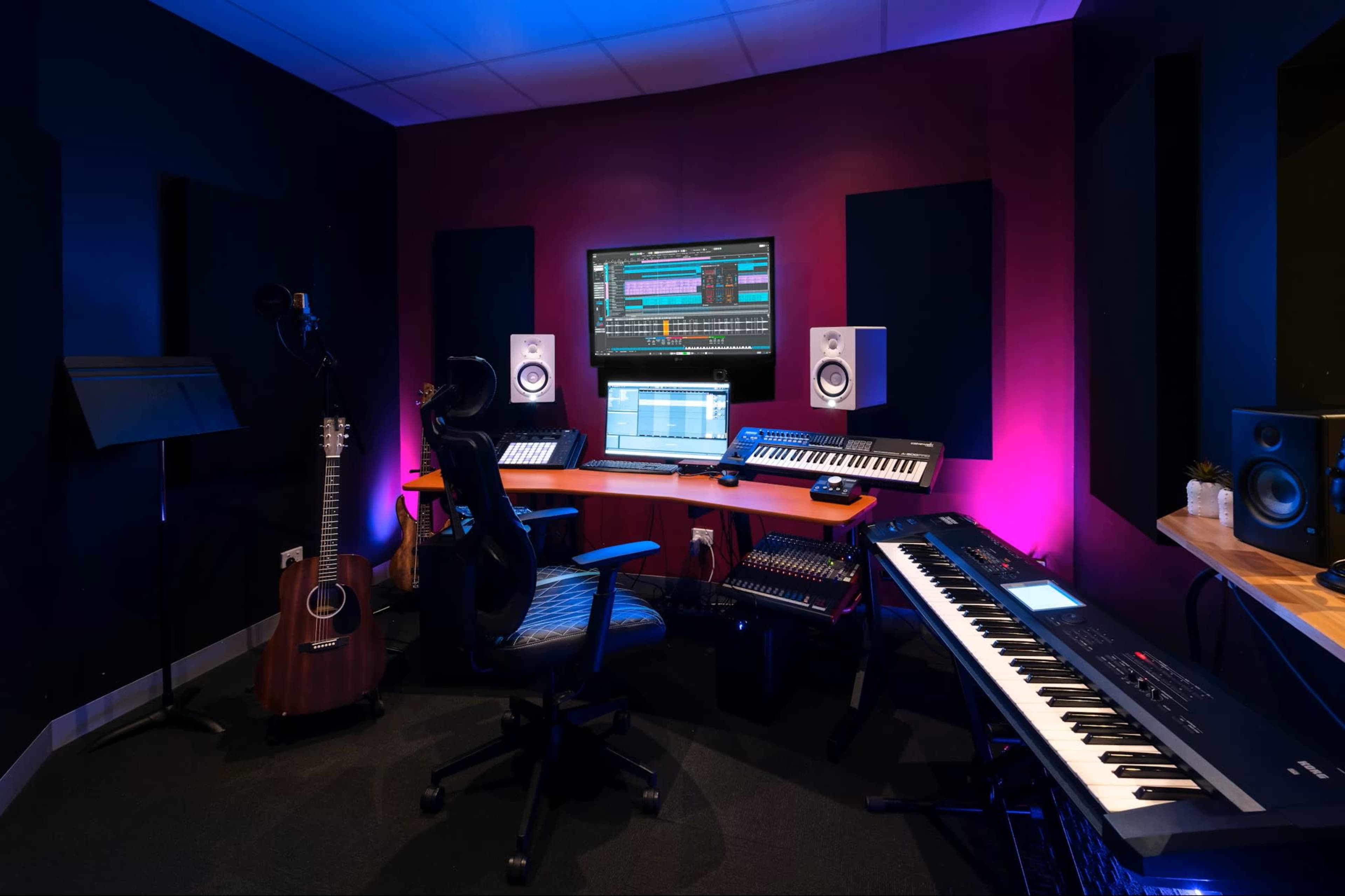The image shows a music production studio featuring a desk with audio equipment, a computer screen displaying software, an acoustic guitar, a keyboard, and ambient lighting.