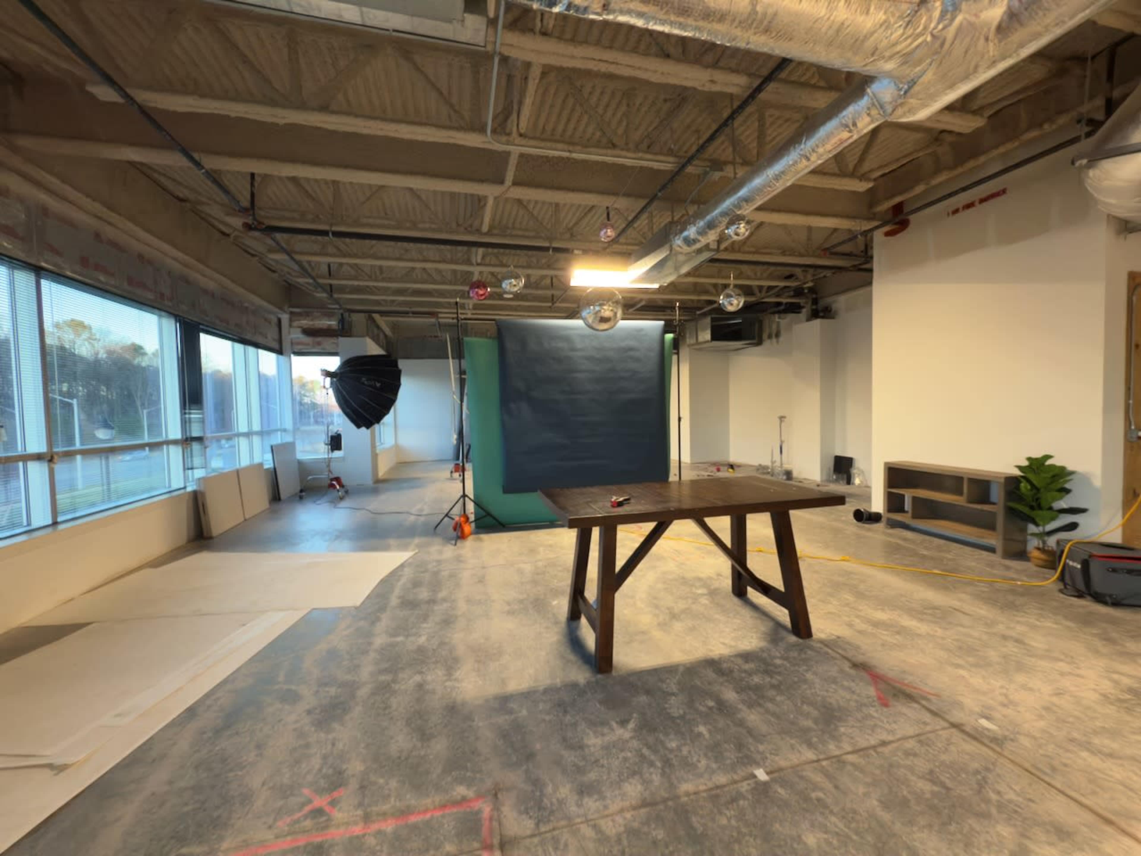 The image shows a sparsely furnished studio space with a wooden table in the center, a green backdrop, and various lighting equipment set up for a photoshoot.