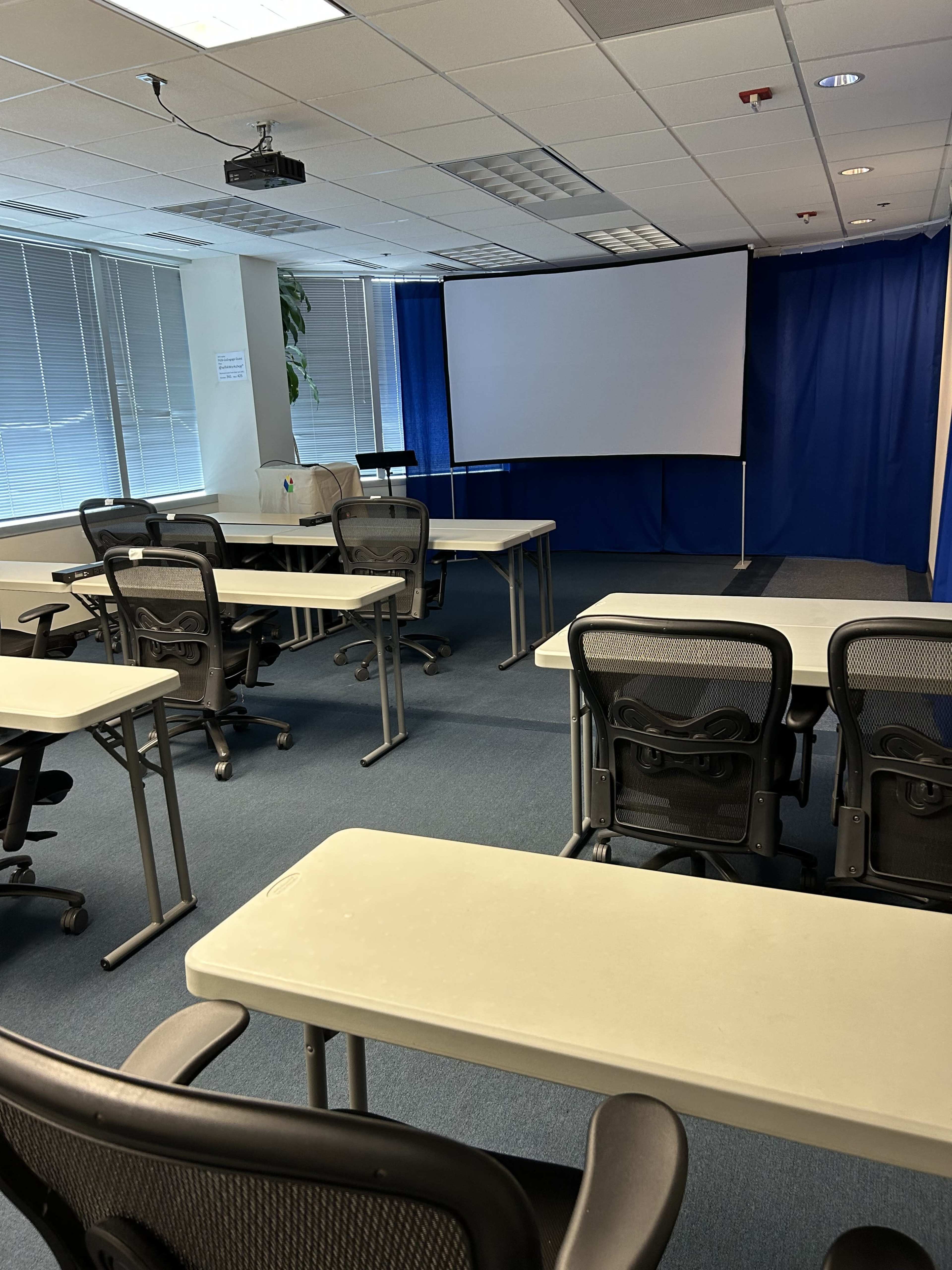 An empty classroom with several tables and chairs arranged facing a large projection screen at the front.