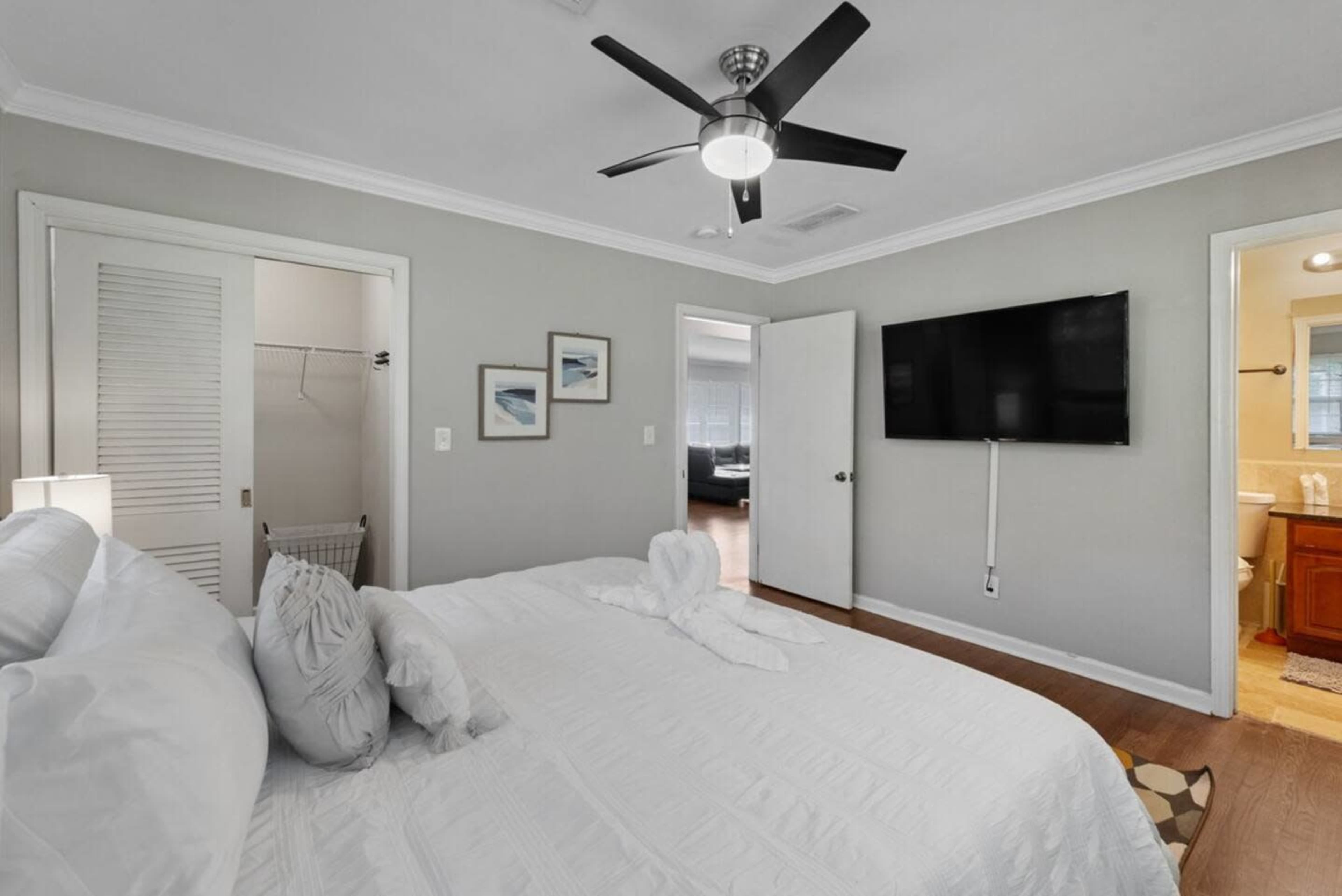 The image shows a well-lit bedroom with a large bed, a ceiling fan, a wall-mounted TV, and an adjoining bathroom visible through an open door.