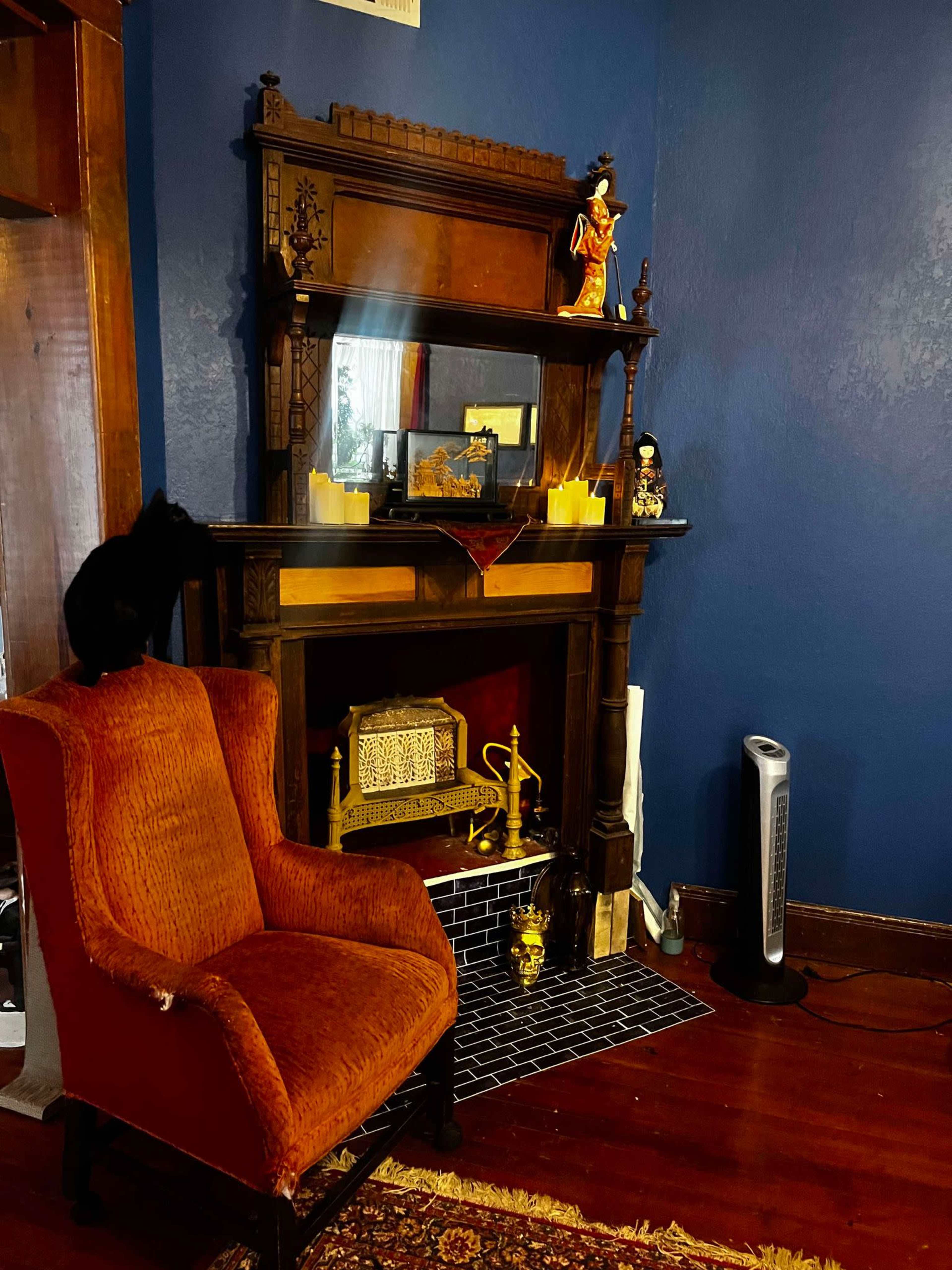 The image features an orange armchair next to a wooden fireplace mantel adorned with decorative items and a small black cat perched on the seat.