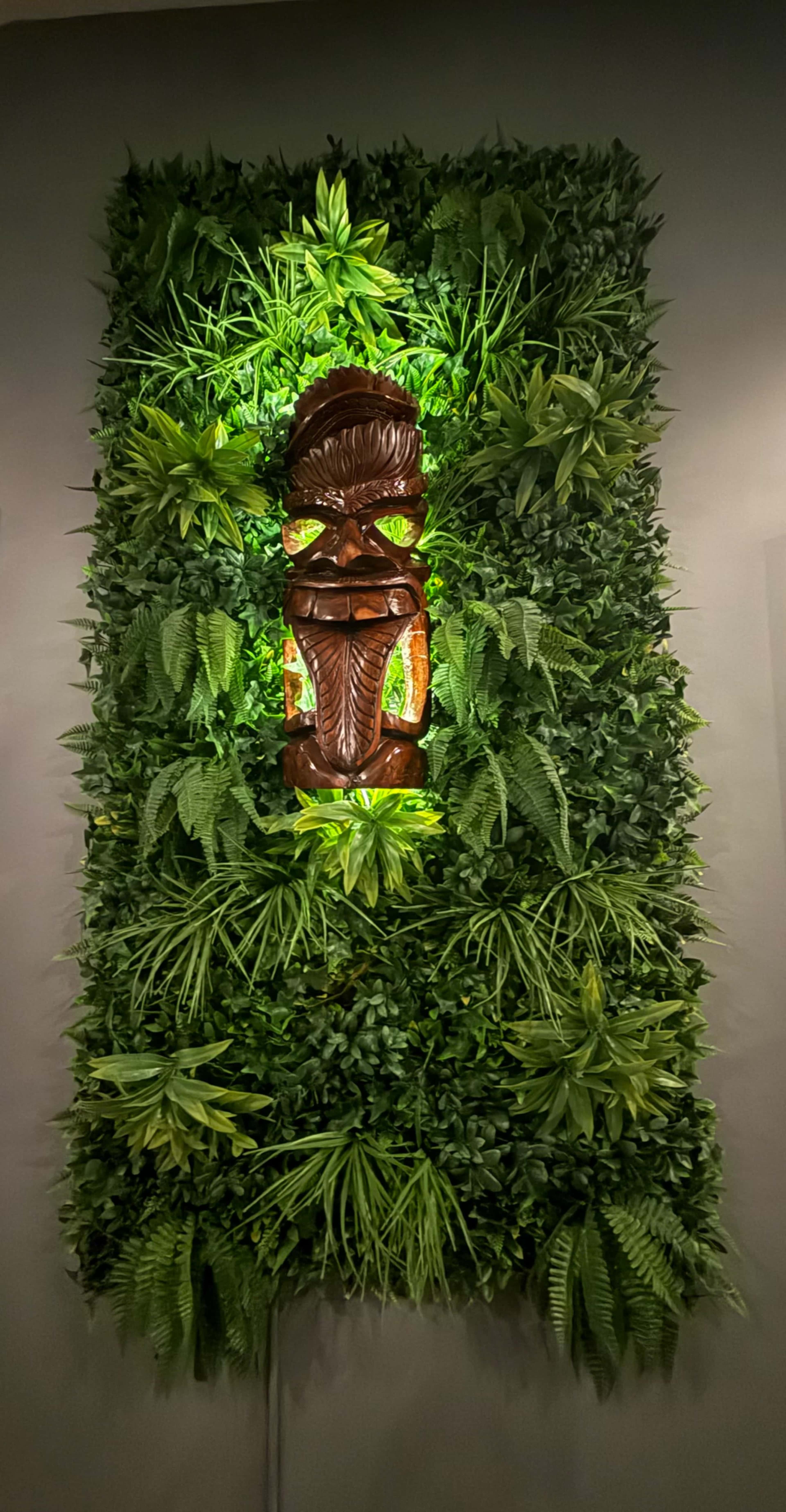 A wooden tiki statue is mounted against a wall covered in green foliage and plants, with soft lighting highlighting the figure.