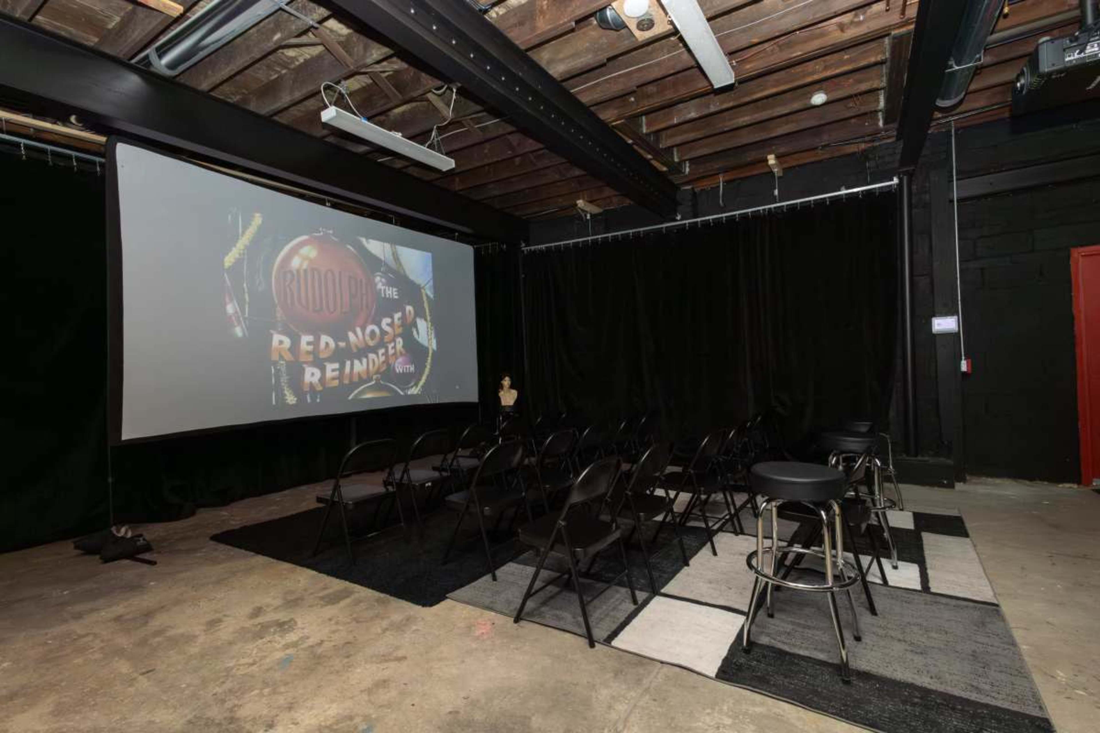 The image shows a dimly lit indoor space with black curtains, a projected screen displaying "Rudolph the Red-Nosed Reindeer," and arranged seating consisting of black chairs and bar stools on a carpeted area.