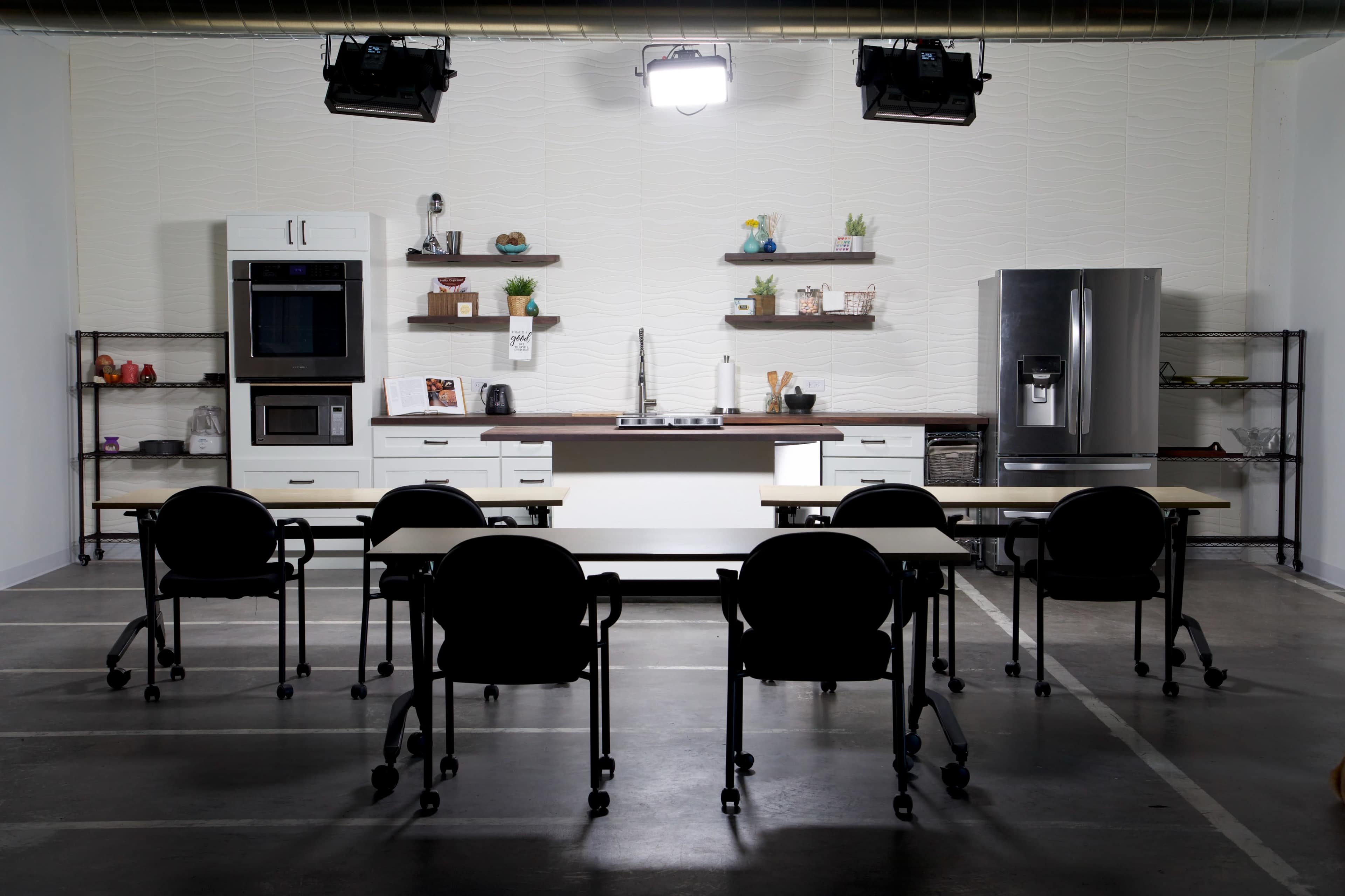 A modern kitchen setup features a stainless steel refrigerator, an oven, and minimalist shelving, with six black rolling chairs positioned around two tables.