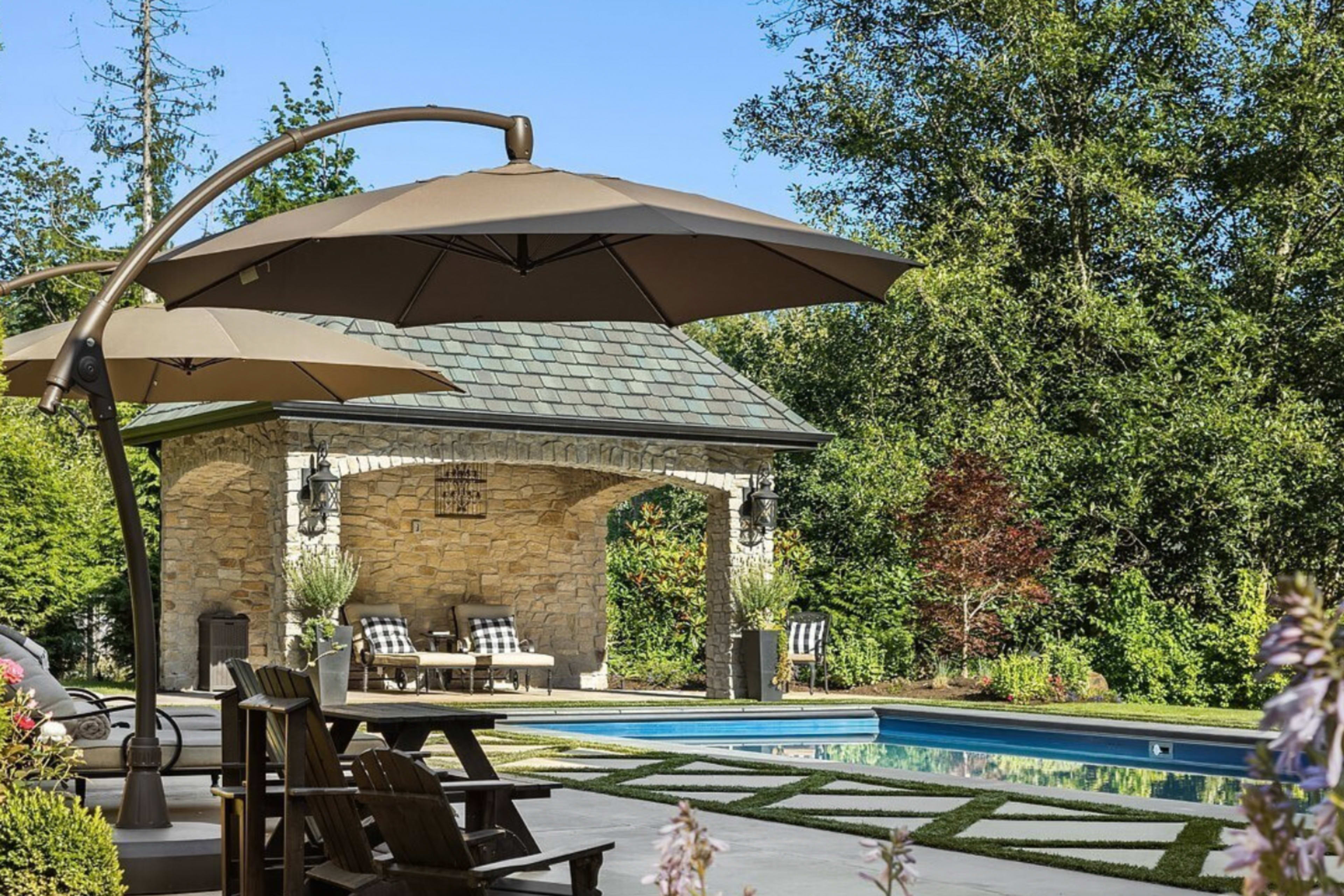 The image shows a landscaped backyard with a swimming pool, a stone pavilion, and shaded seating areas under large umbrellas.