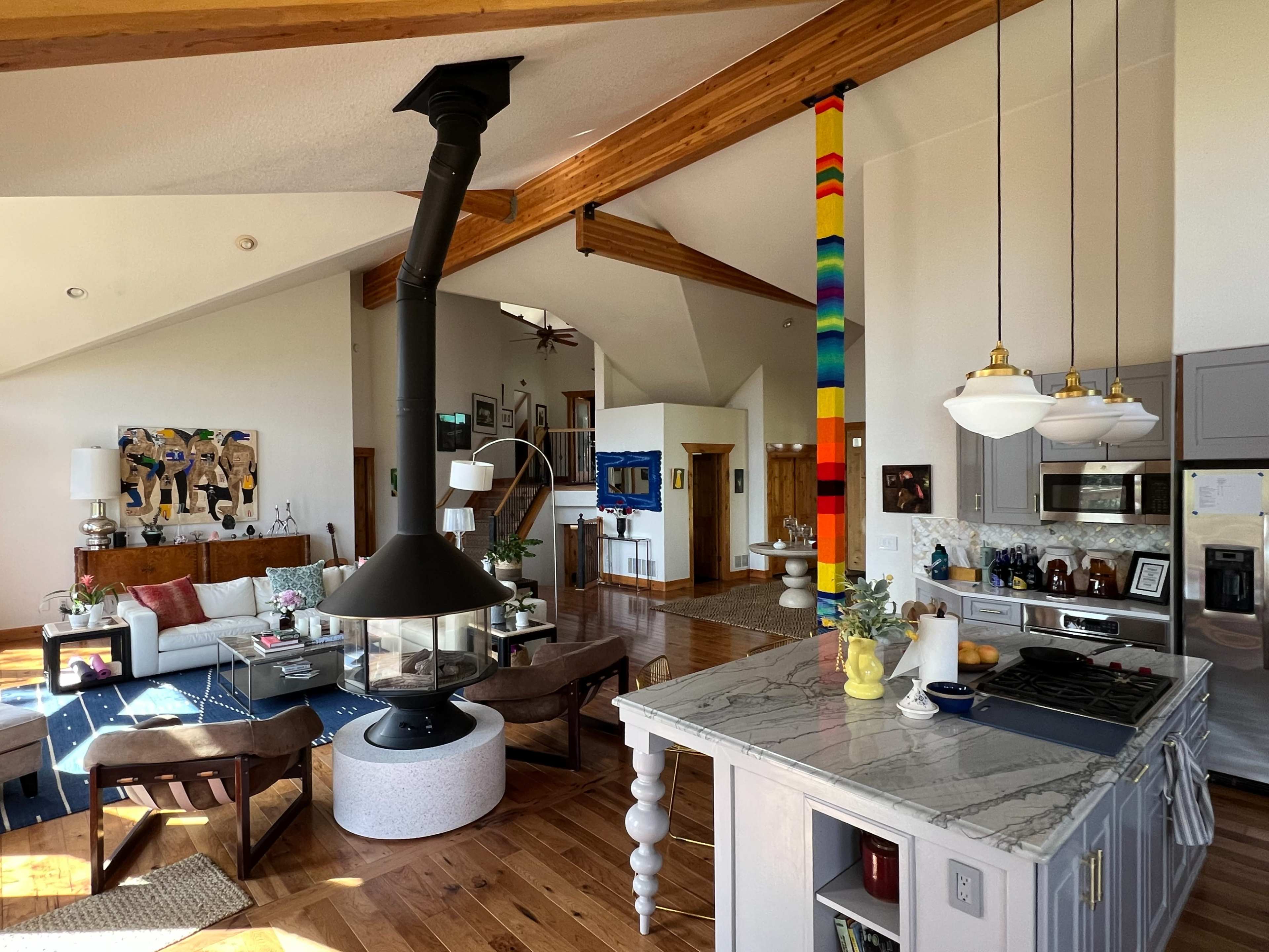 The image shows an open-concept living space featuring a kitchen, dining area, and a living room with wooden beams, modern furnishings, and a colorful art installation.
