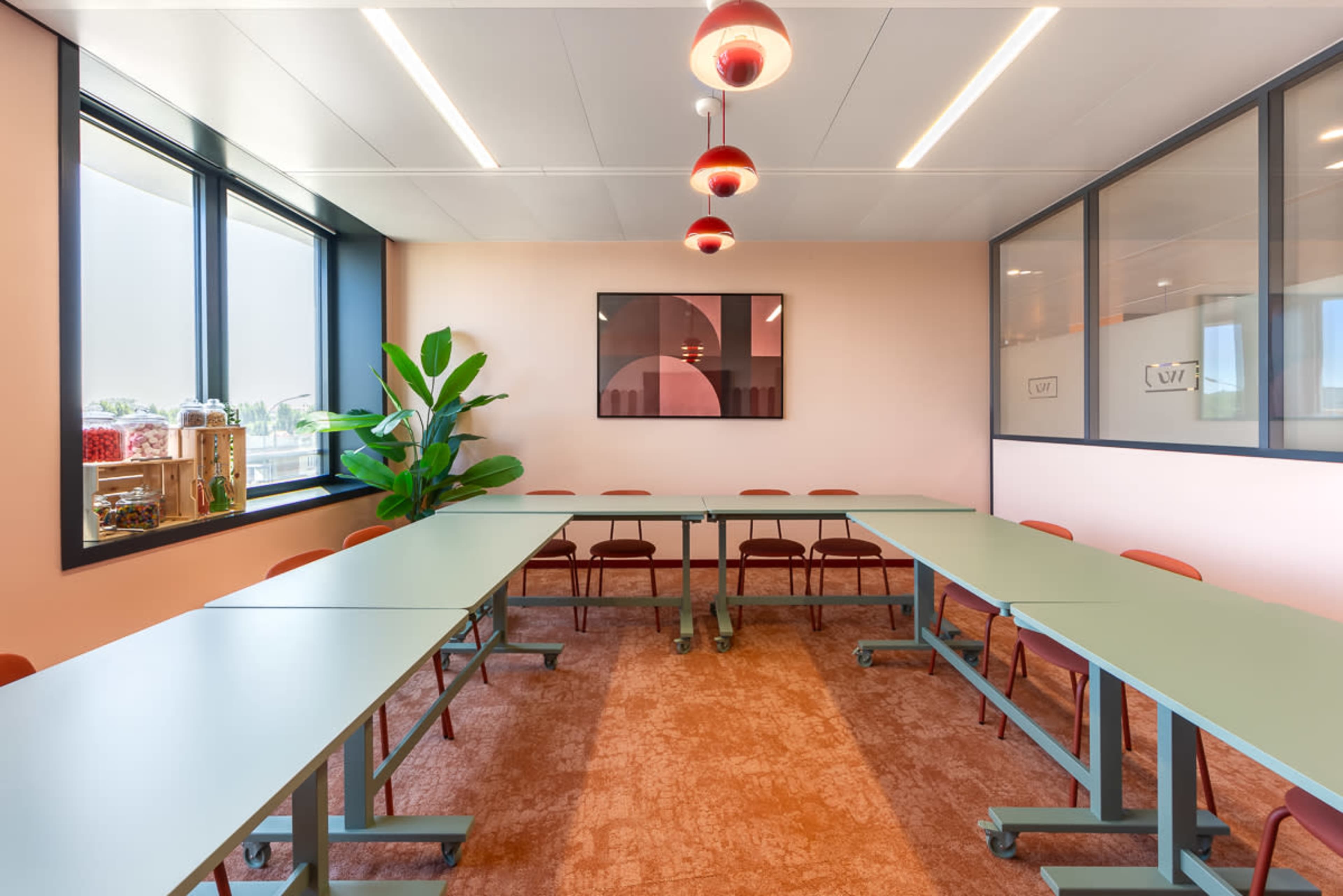 The image shows a modern conference room with a long, green table, red chairs, and large windows that provide natural light.