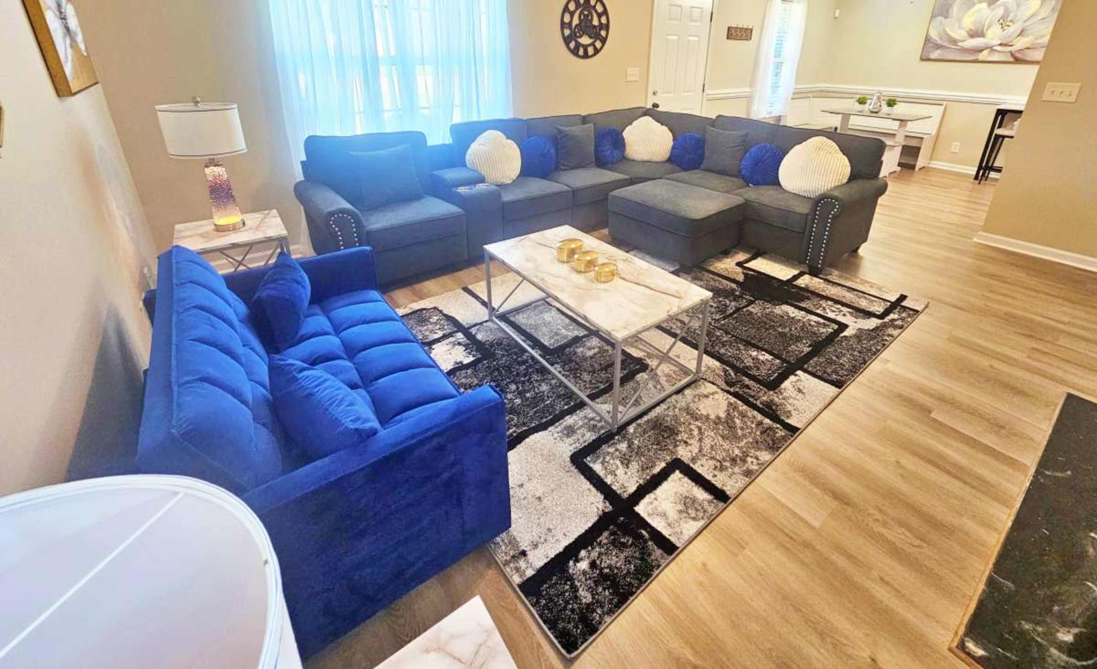 A spacious living room features a large blue sectional sofa, a matching loveseat, and a coffee table set on a patterned rug under soft natural light from a nearby window.