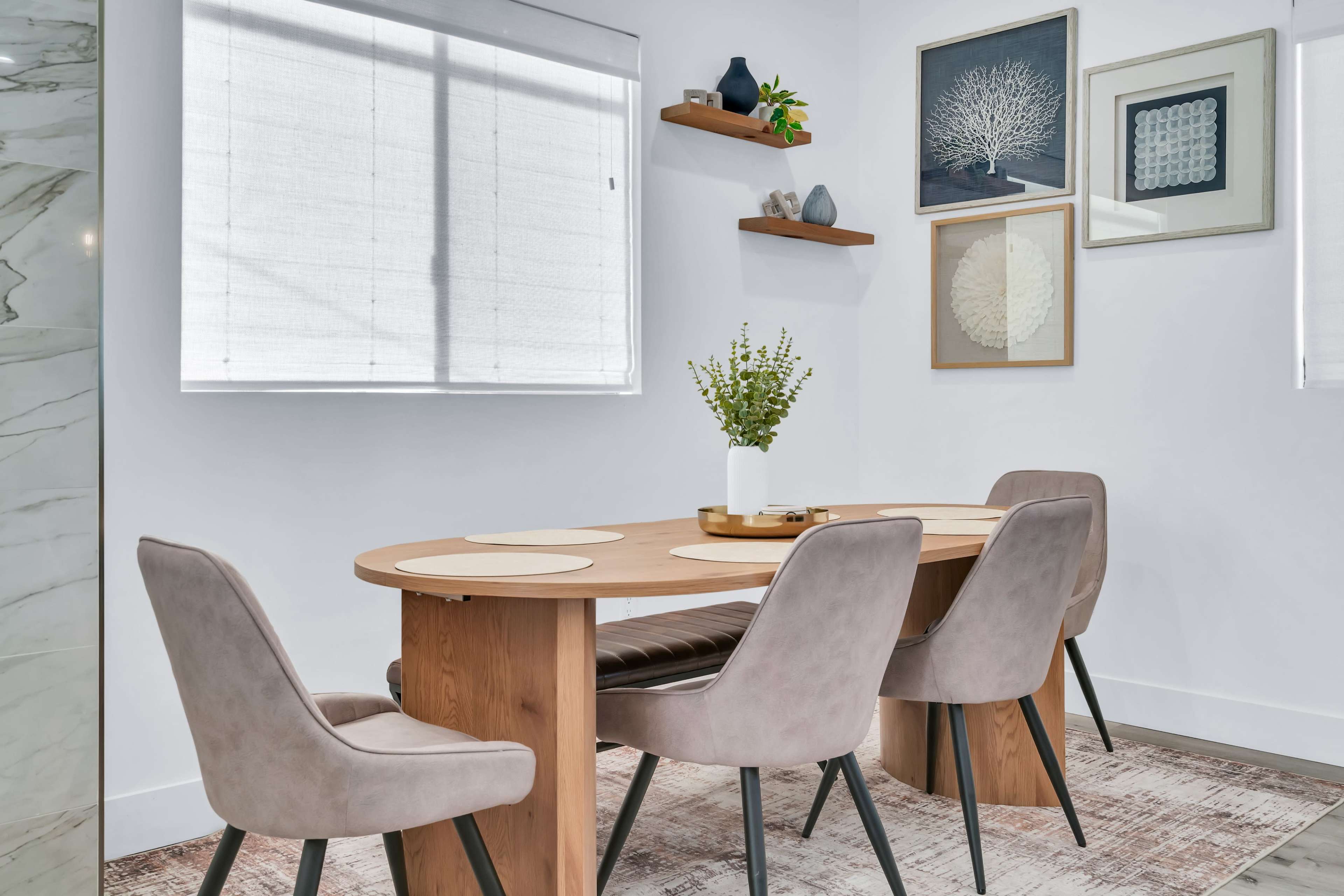 The image shows a modern dining area featuring a wooden table surrounded by four upholstered chairs, with wall shelves displaying decorative items and framed artwork.