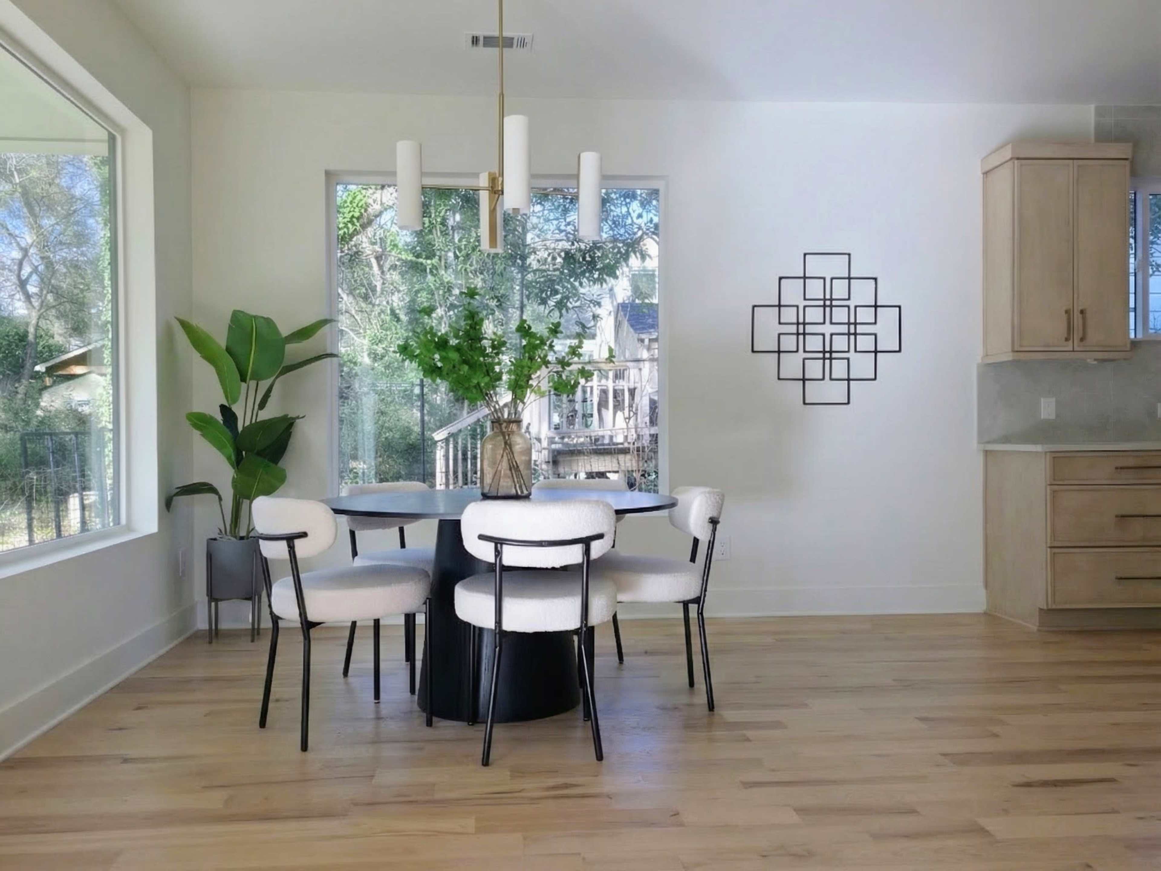 A modern dining area features a round black table surrounded by four upholstered chairs, with large windows allowing natural light and a view of greenery outside.