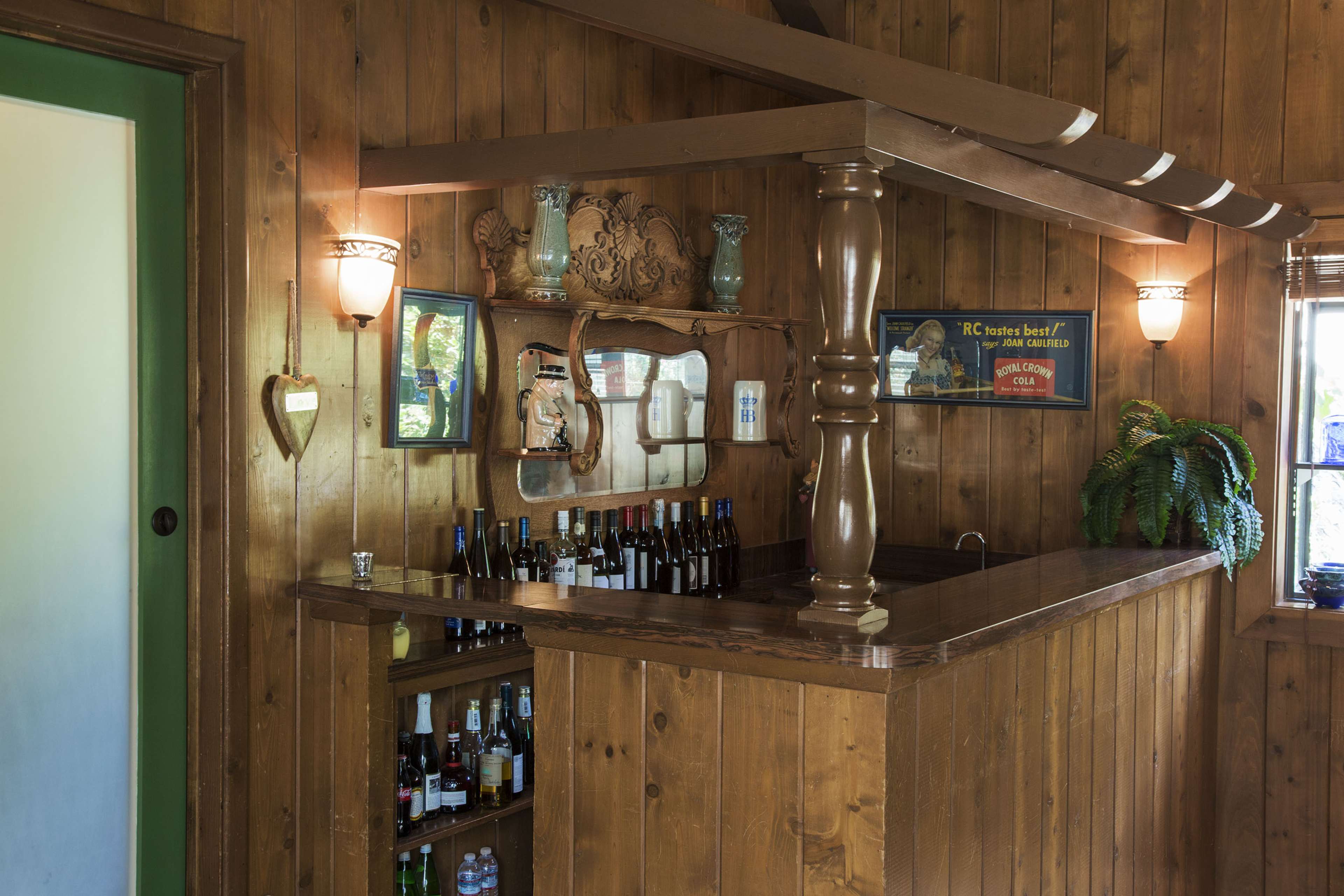 The image shows a wooden bar area with various bottles displayed on shelves and a mirror above the bar, surrounded by rustic wood paneling.