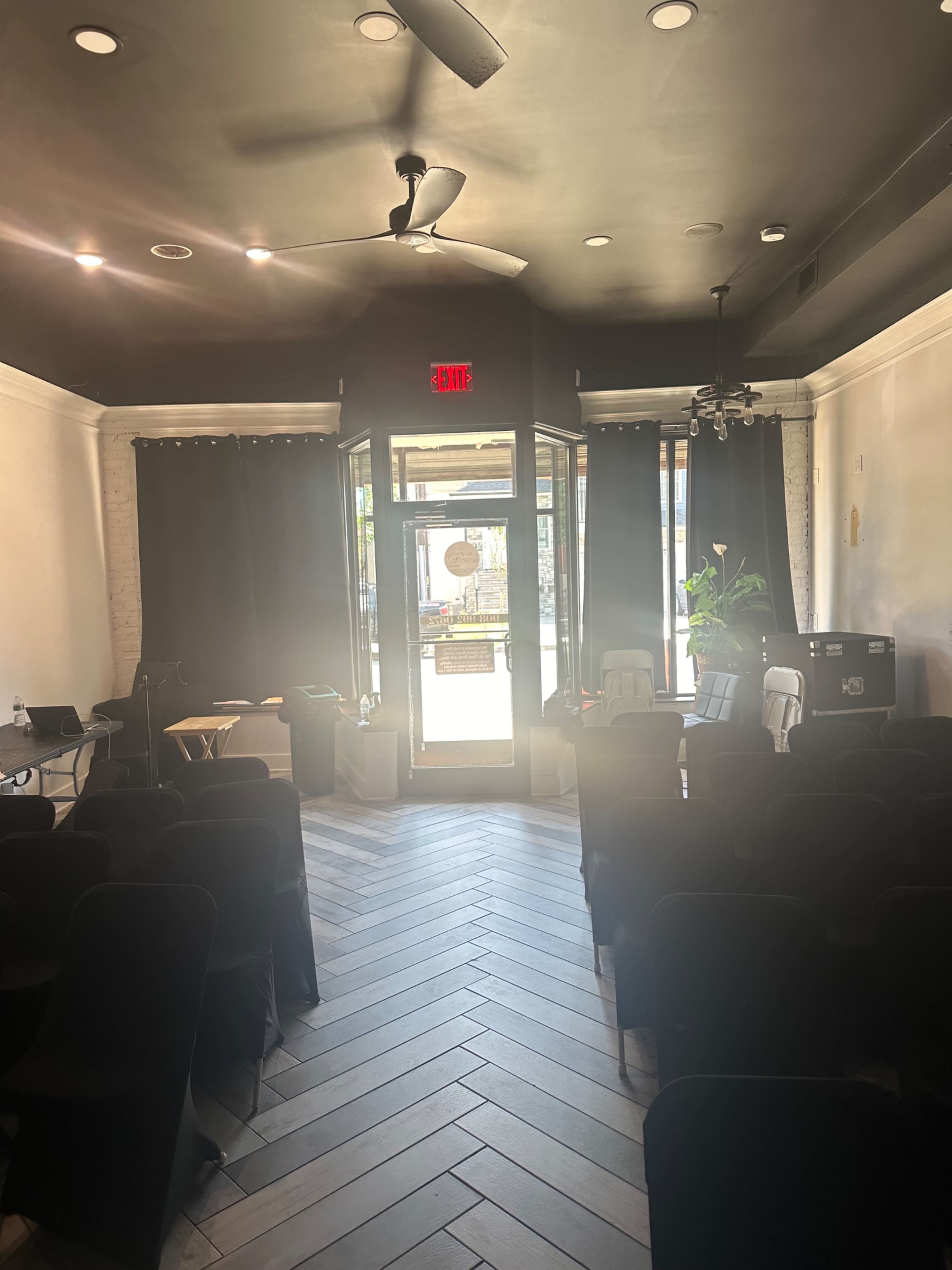 An empty room with rows of black chairs facing a doorway, illuminated by natural light, and featuring large windows covered with dark curtains.