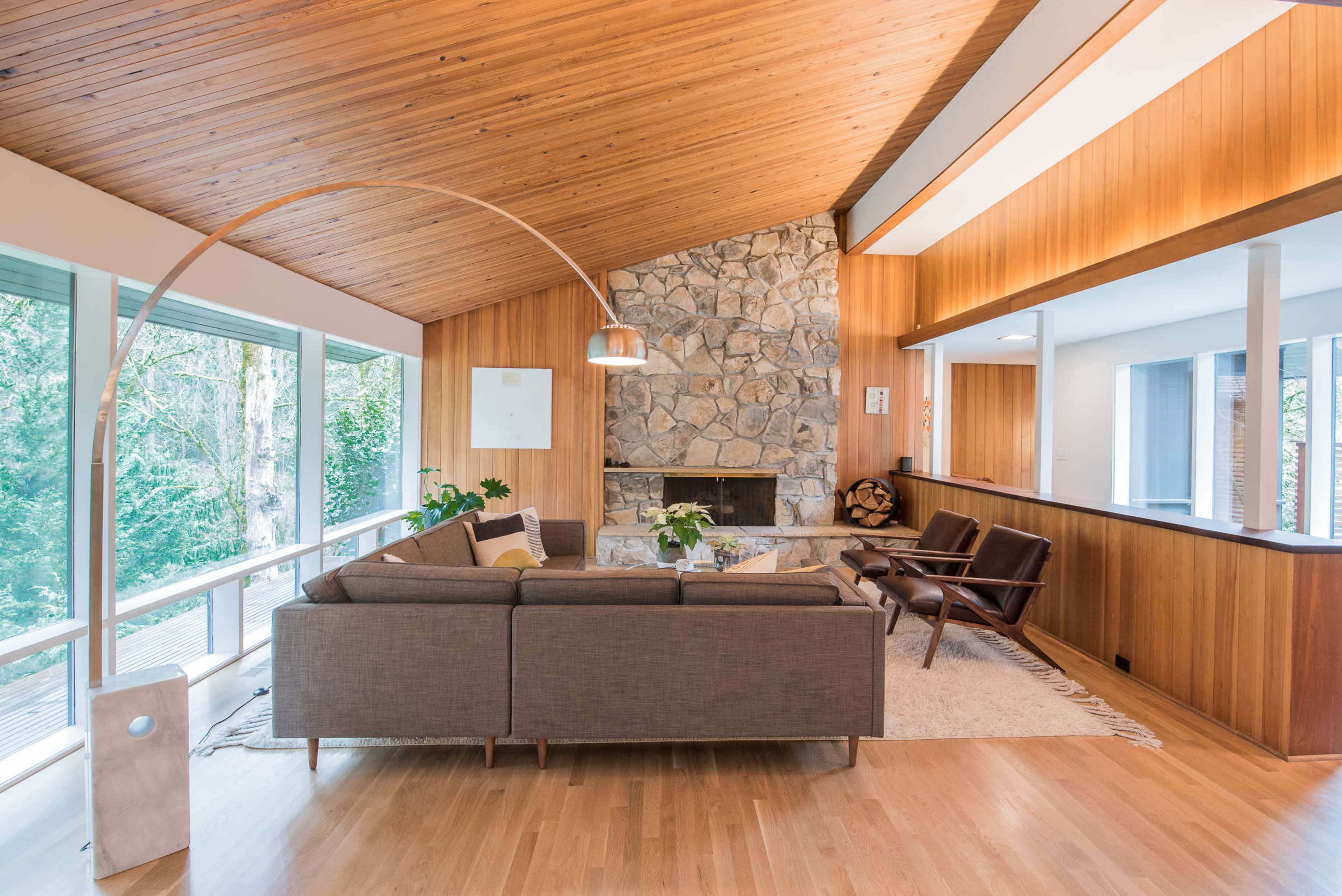 The living room features a stone fireplace, a curved floor lamp, and large windows that offer views of the surrounding greenery.