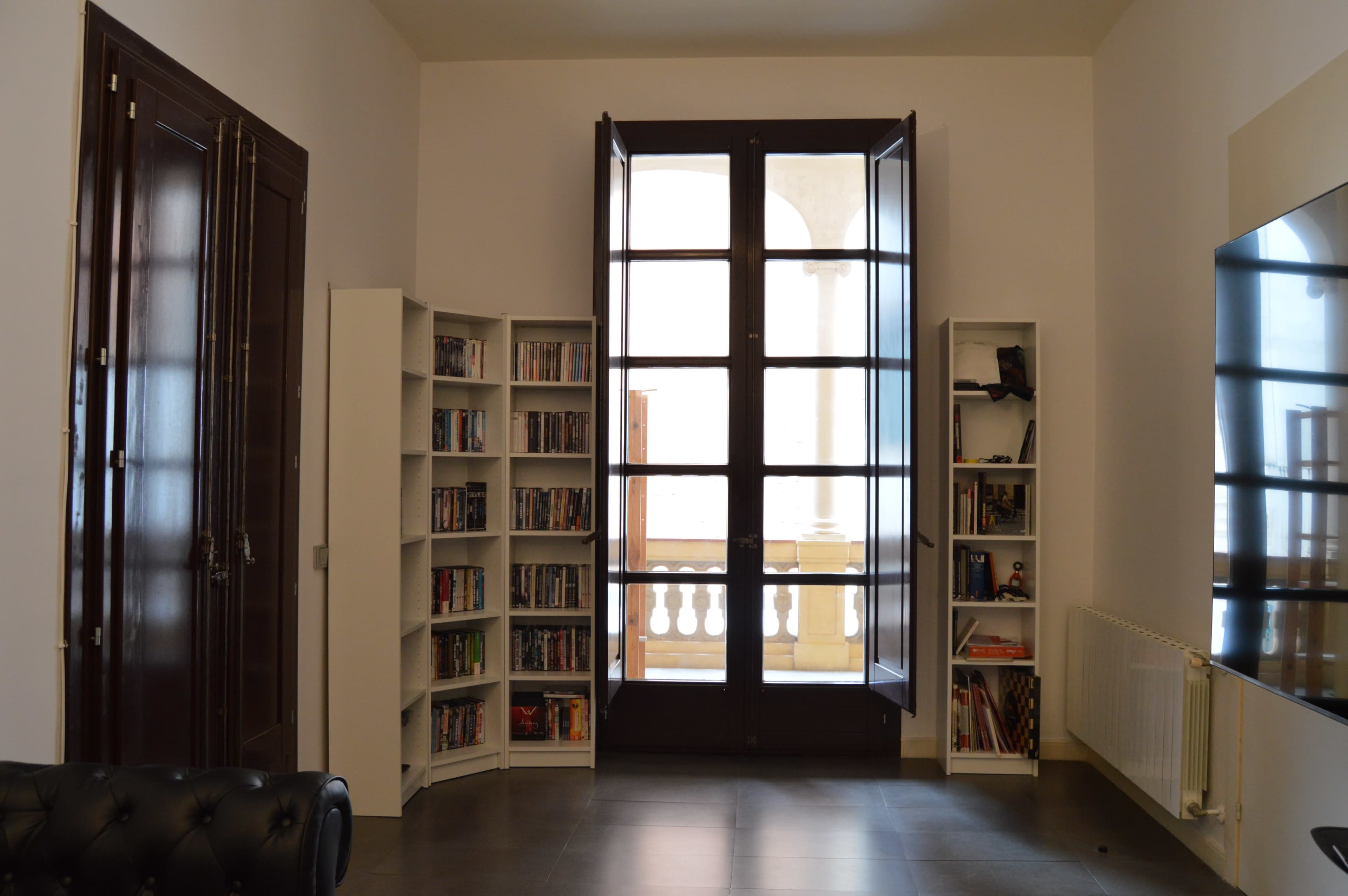 A room with tall windows, a black leather sofa, and two white bookshelves filled with books and media.