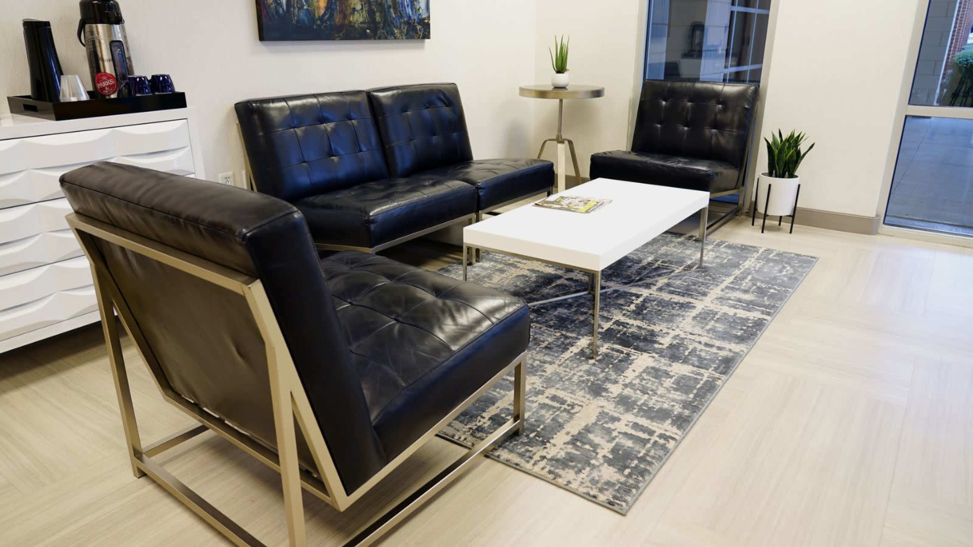 A modern waiting area features a black leather sofa, two matching chairs, a white coffee table, and a decorative plant, all arranged on a patterned rug.