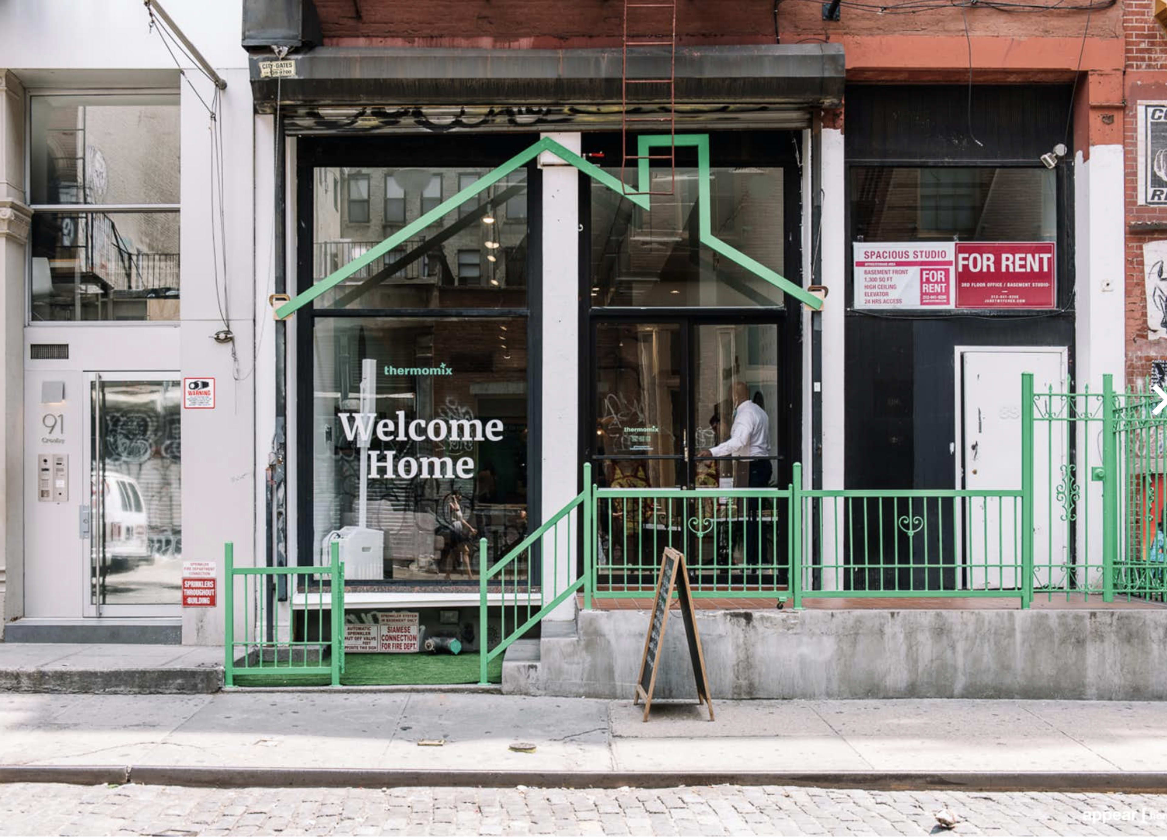 A storefront with large windows displaying a "Welcome Home" sign, and a green outline of a house, alongside a "For Rent" sign and a fenced area.