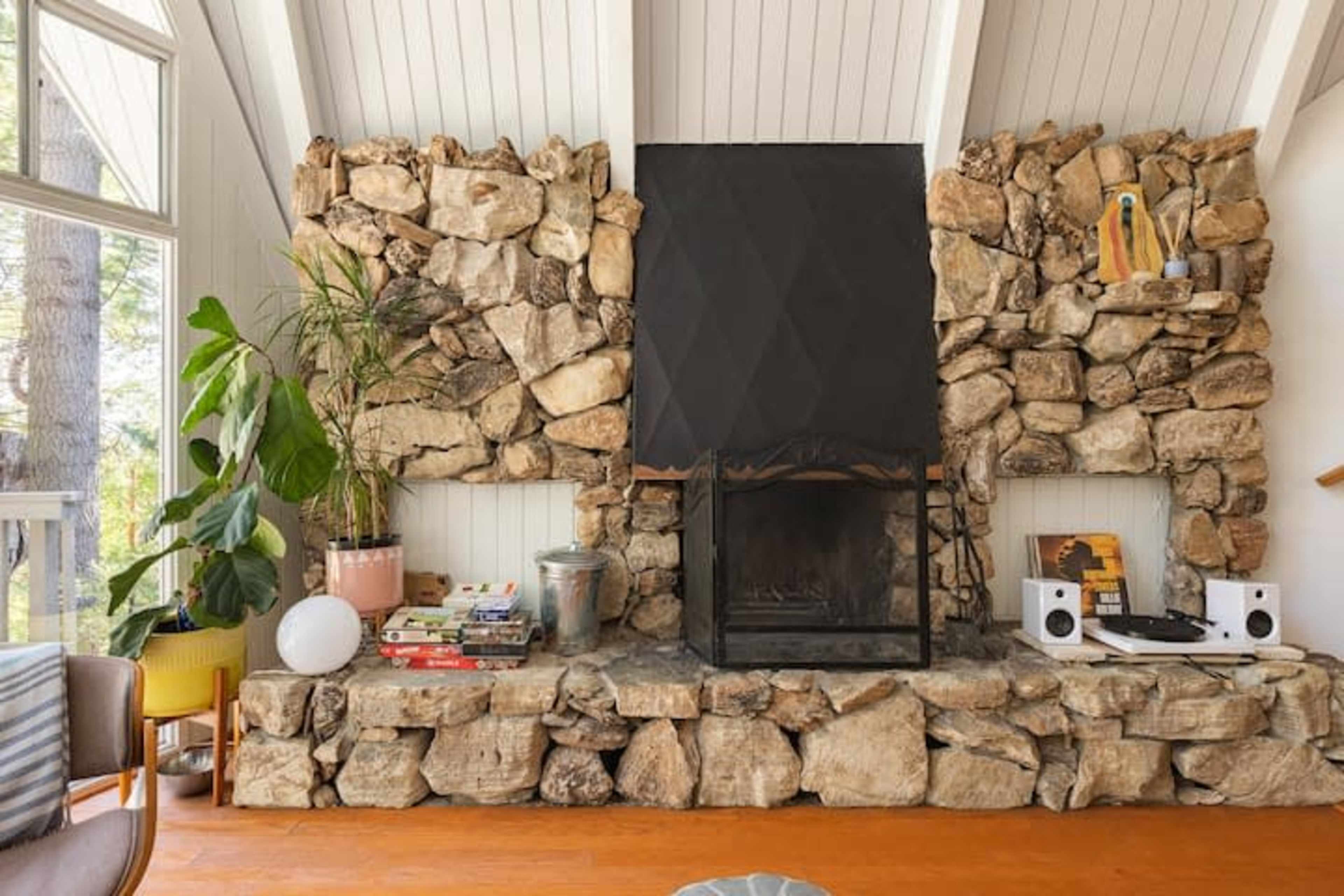 A living room with a large stone fireplace, surrounded by white walls and furniture, featuring plants and decorative items nearby.