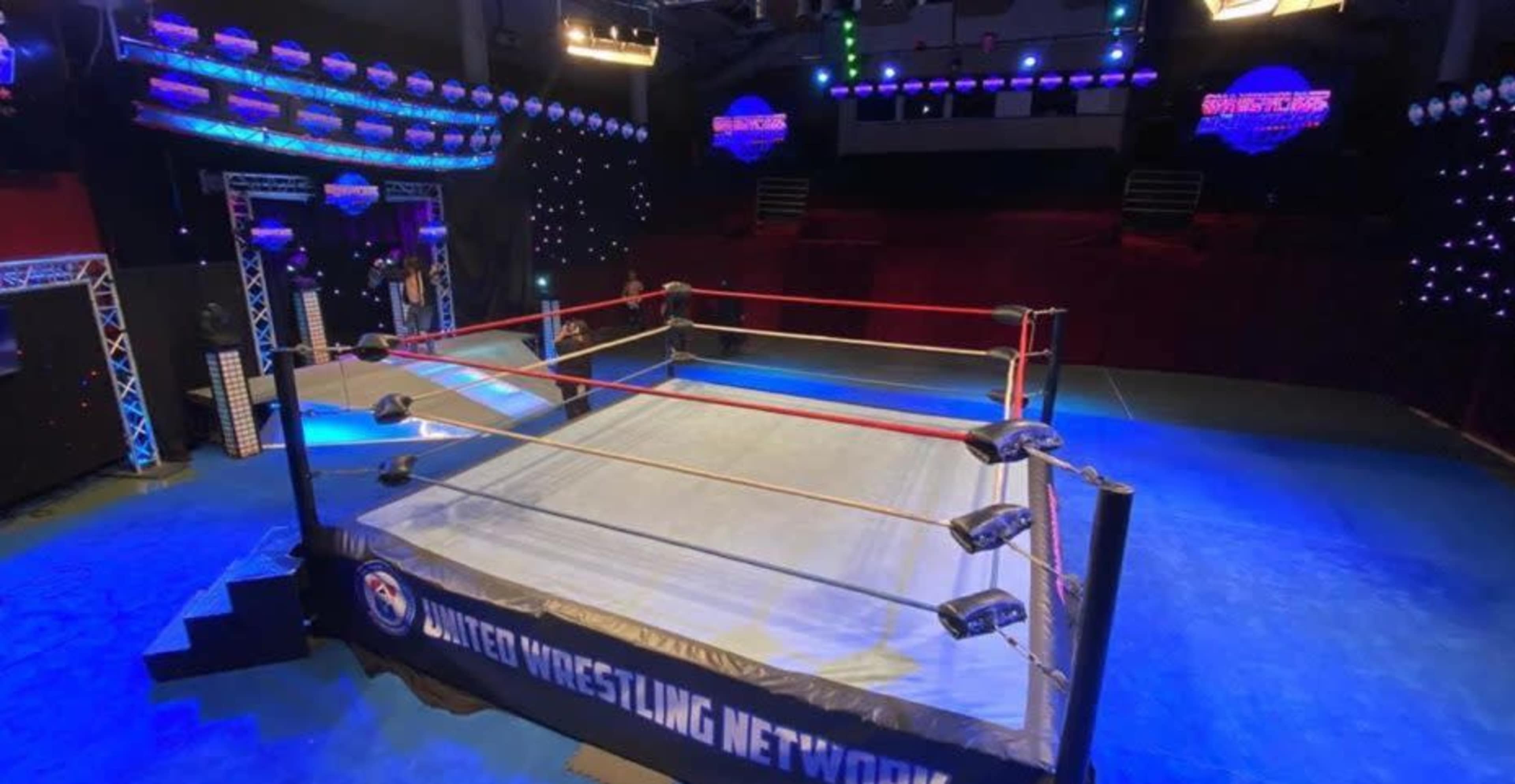 The image shows a wrestling ring in a brightly lit arena, featuring red ropes and a blue mat, with banners for the United Wrestling Network.
