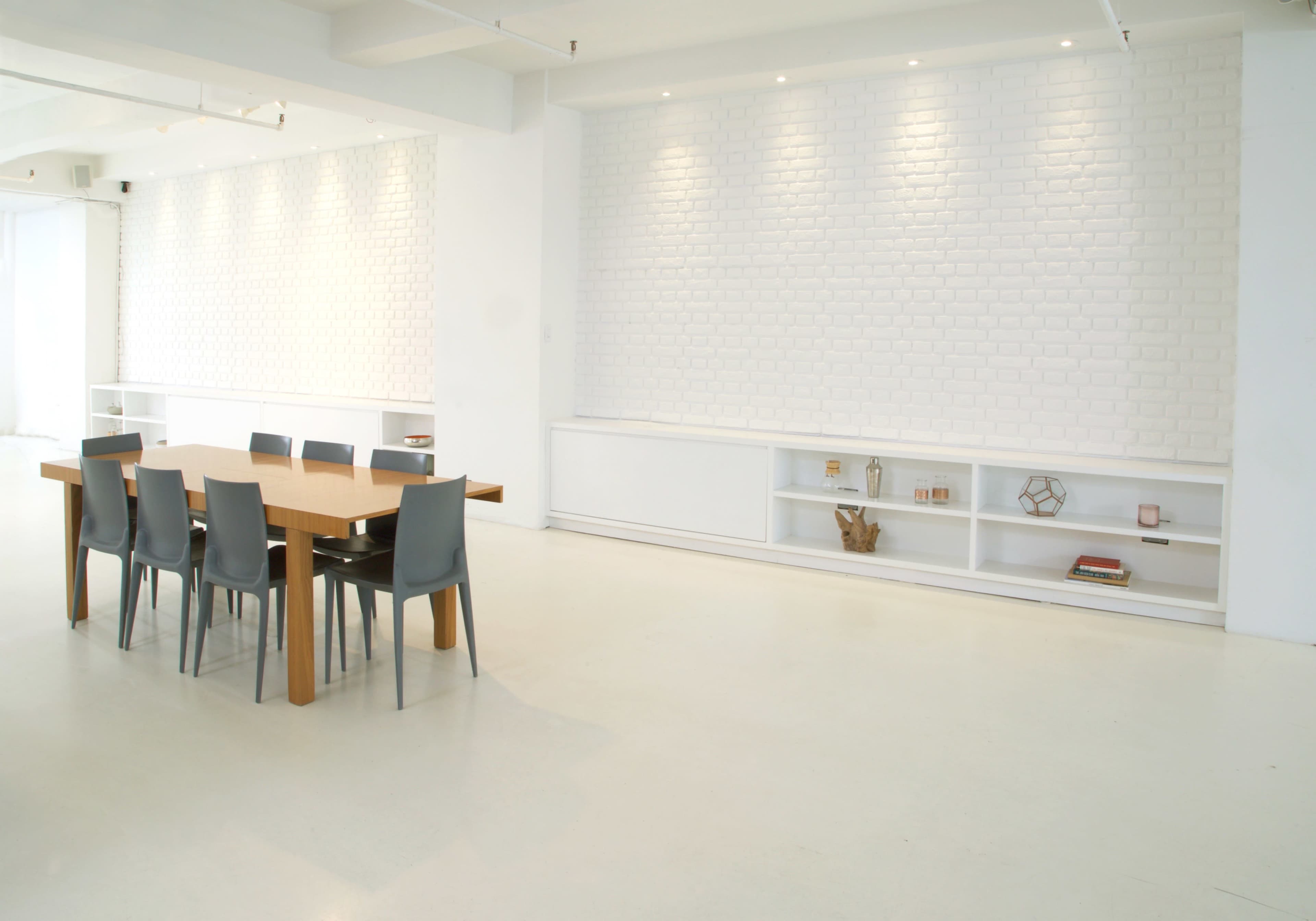 A modern dining area features a wooden table surrounded by gray chairs, set against a white brick wall and minimal shelving.