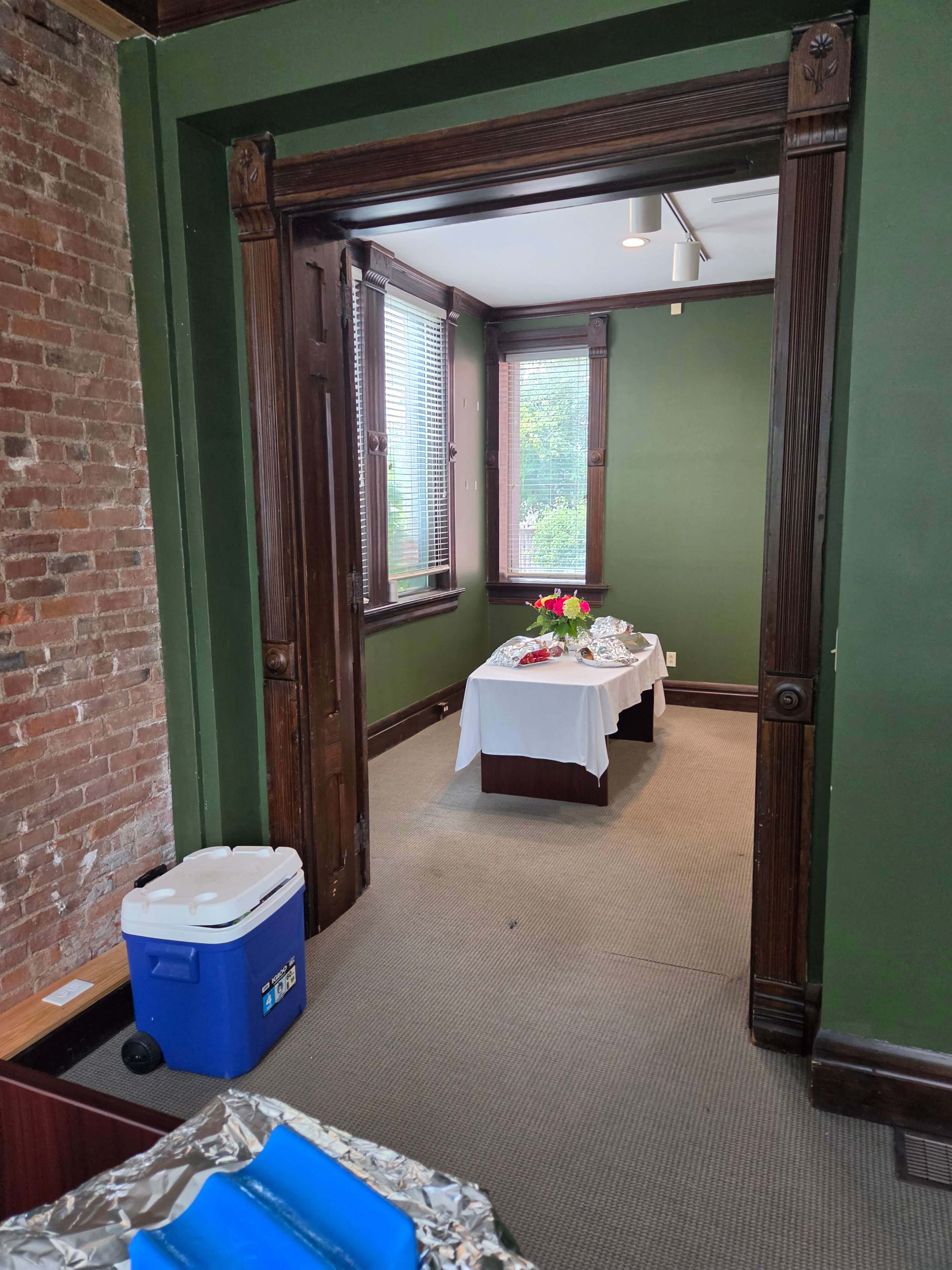 A neatly arranged table with flowers is set up in a room featuring exposed brick walls and green paint, visible through an open doorway.