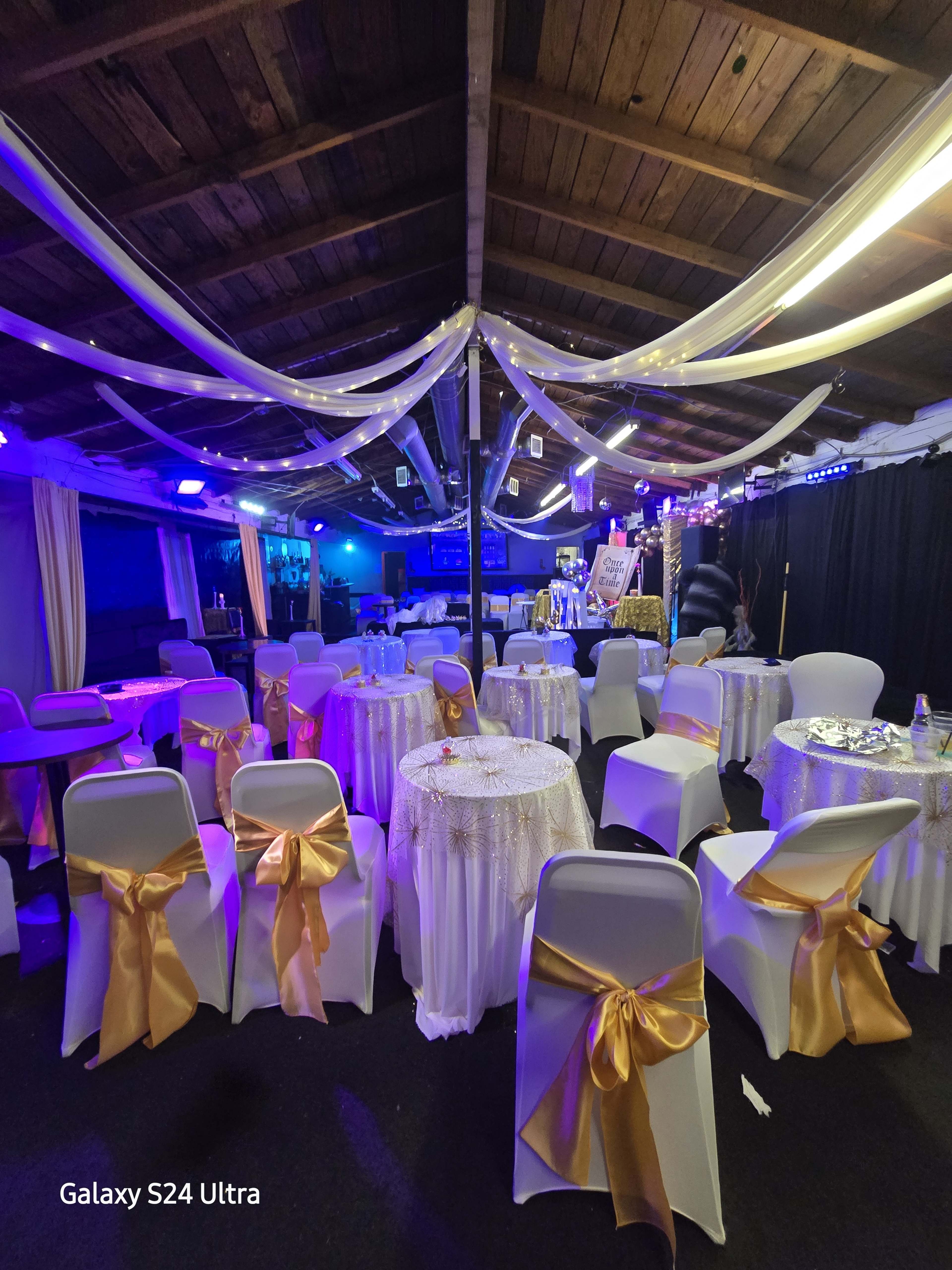 The image shows a decorated event space with tables dressed in white tablecloths and gold sashes, under soft draped lighting.