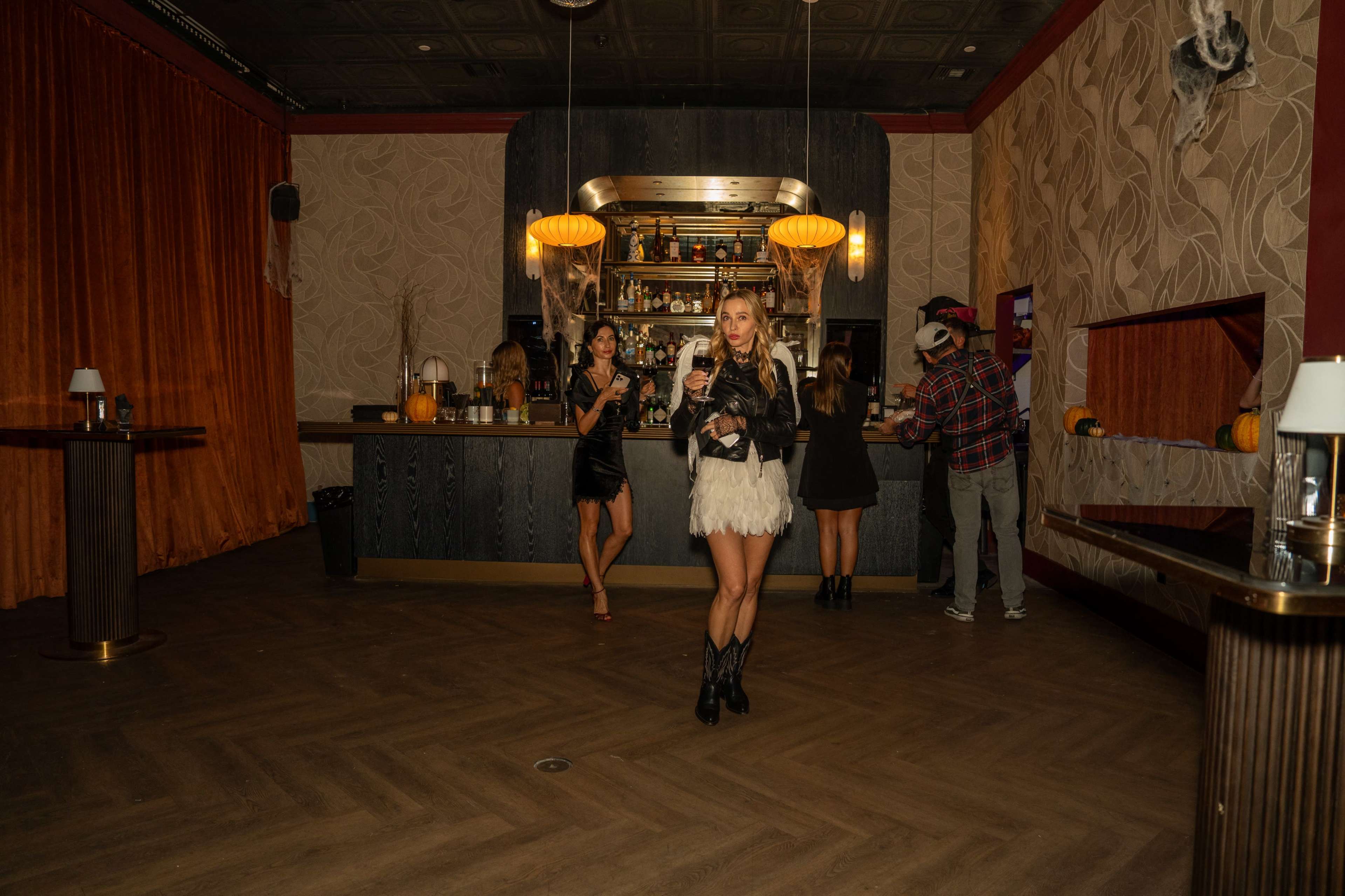 A group of people gather in a dimly lit bar adorned with autumn decorations, while one woman stands in the foreground holding a drink.