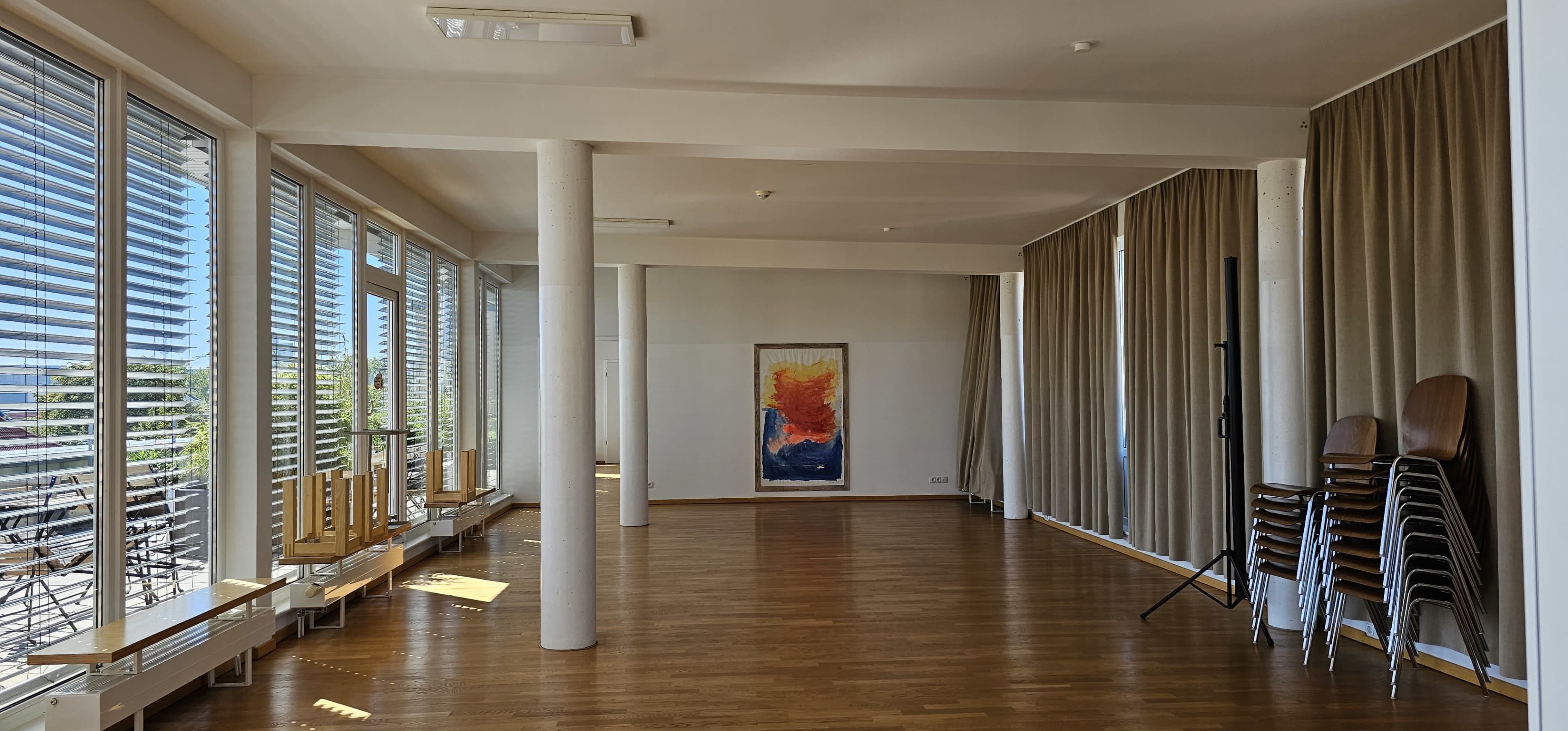 The image shows a spacious interior room with wooden flooring, large windows, and a colorful painting on the wall, alongside stacked chairs and curtains.