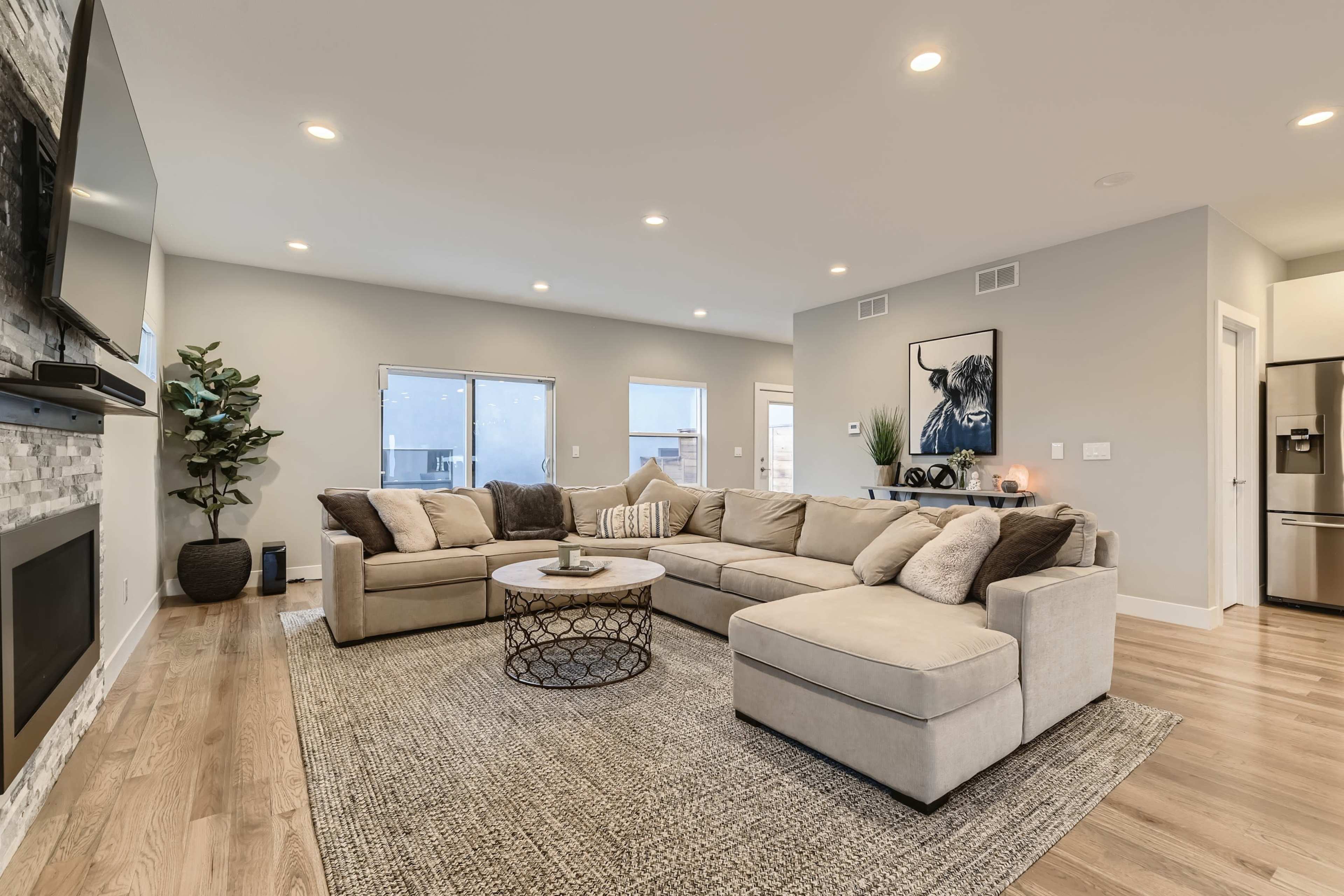 A spacious living room features a large sectional sofa, a round coffee table, and a media console with a television, all complemented by natural light from multiple windows.