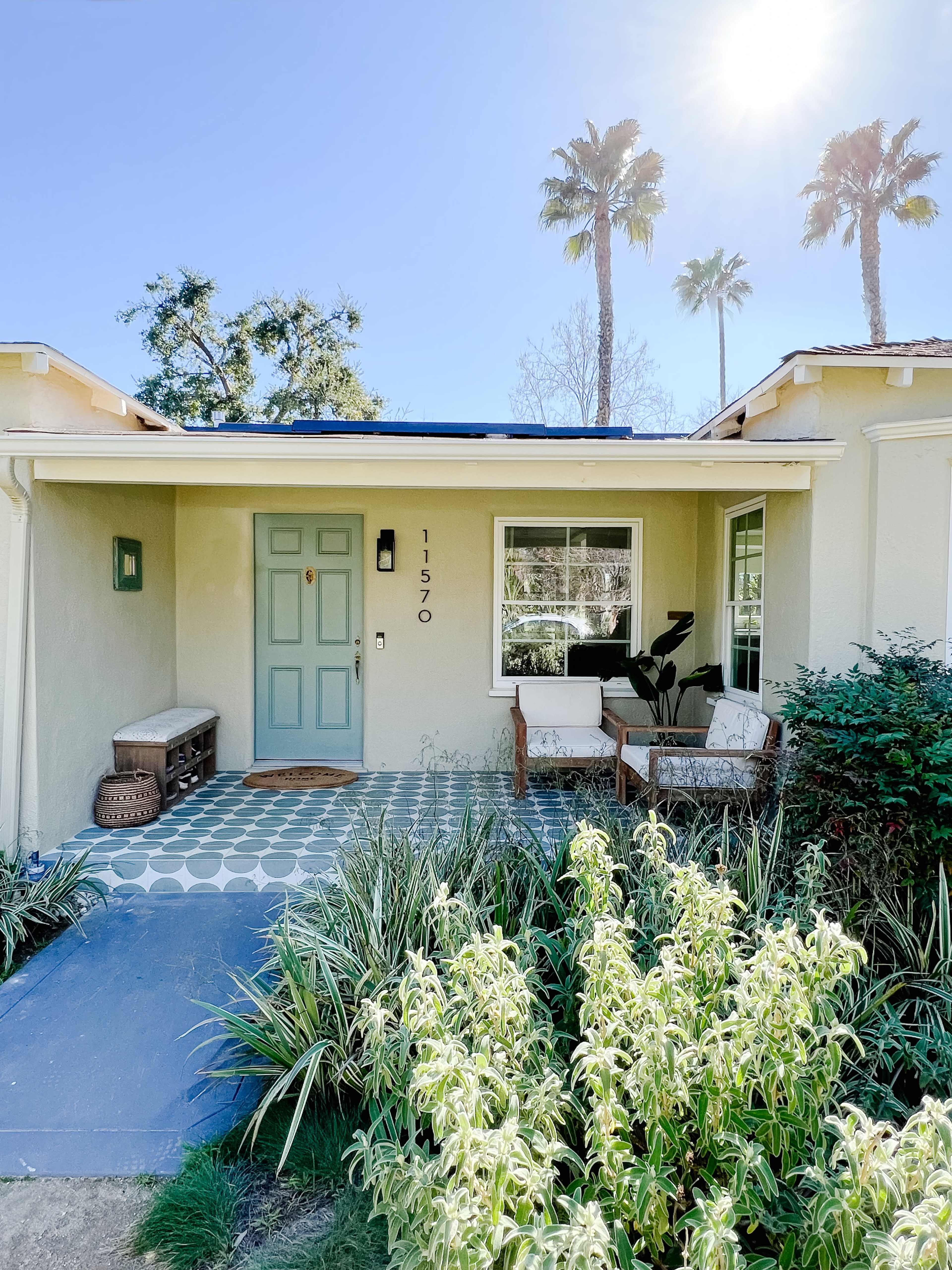 A house entrance with a light blue door, a bench, and a small seating area surrounded by green plants and palm trees.
