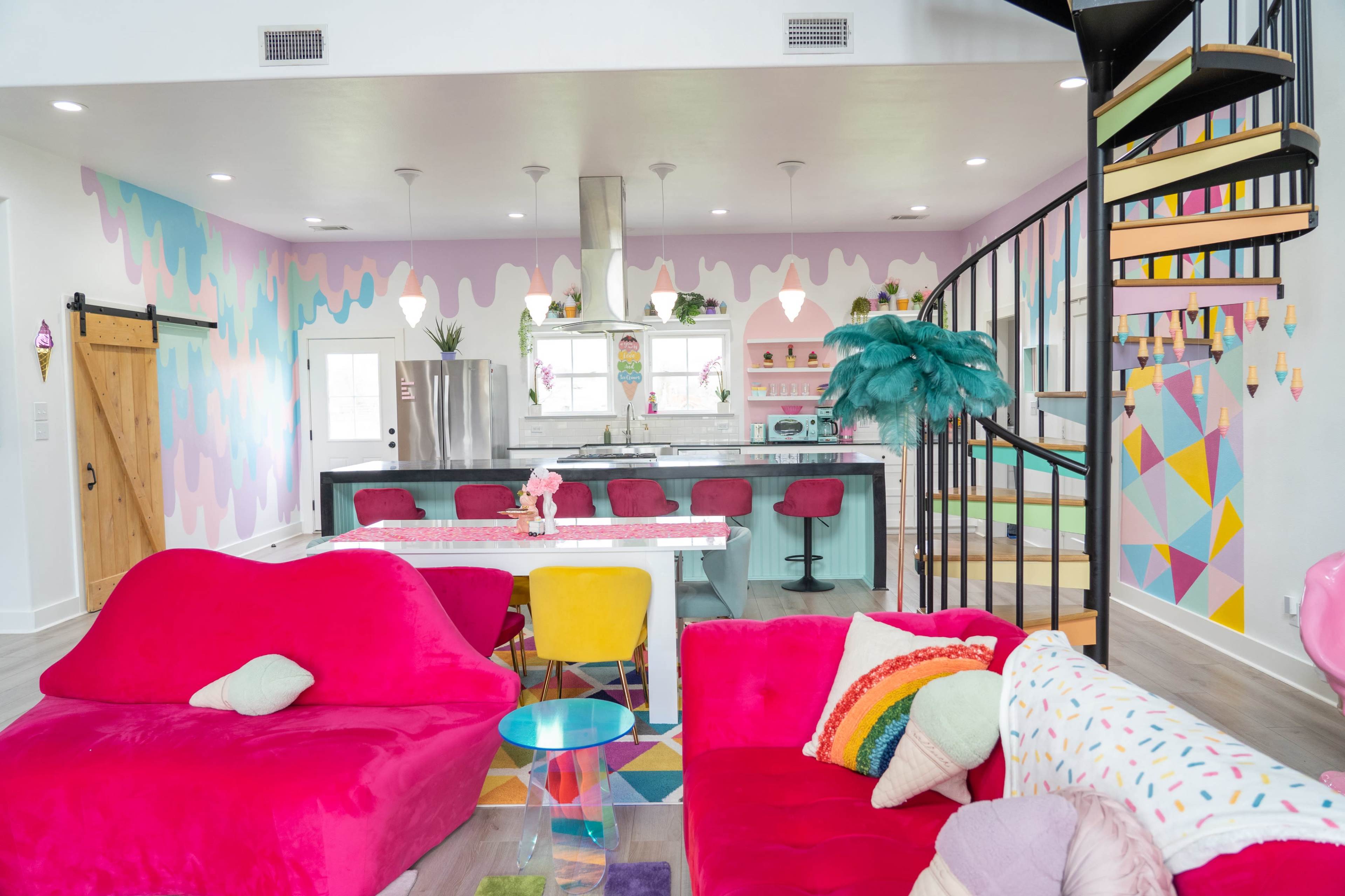 A colorful modern living space features vibrant pink and yellow furniture, a spiral staircase, and a kitchen with a patterned wall and island seating.