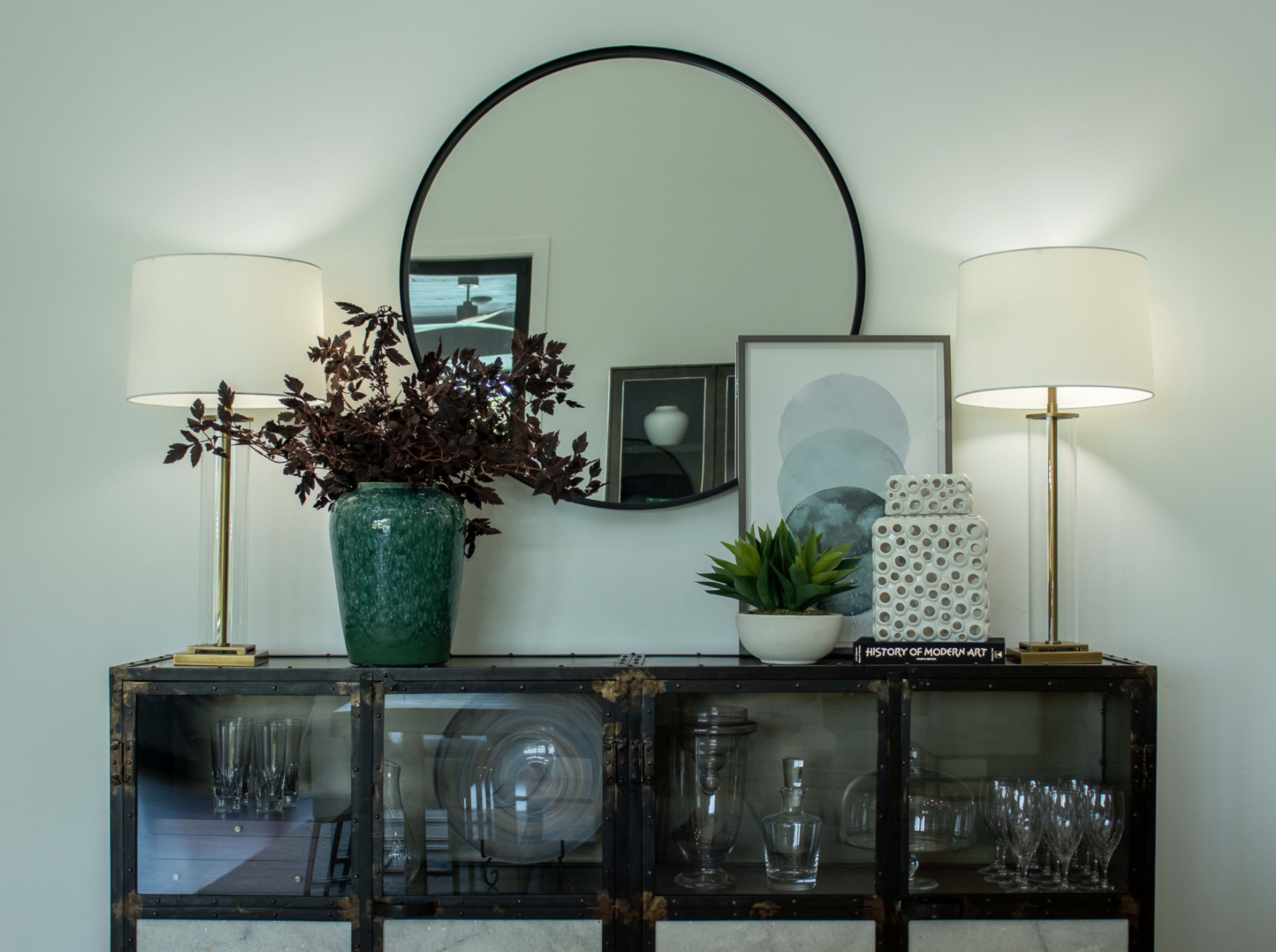 A wooden sideboard holds a green ceramic vase with dried branches, a small potted plant, and a decorative white object, flanked by two lamps and a round mirror on a white wall.