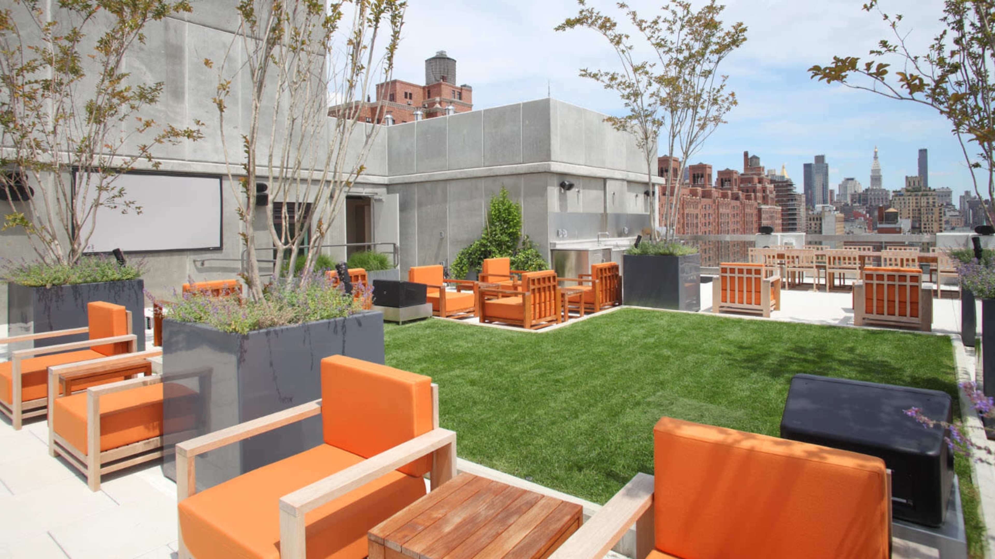 A rooftop patio featuring orange chairs, wooden tables, and potted trees, overlooking a city skyline.