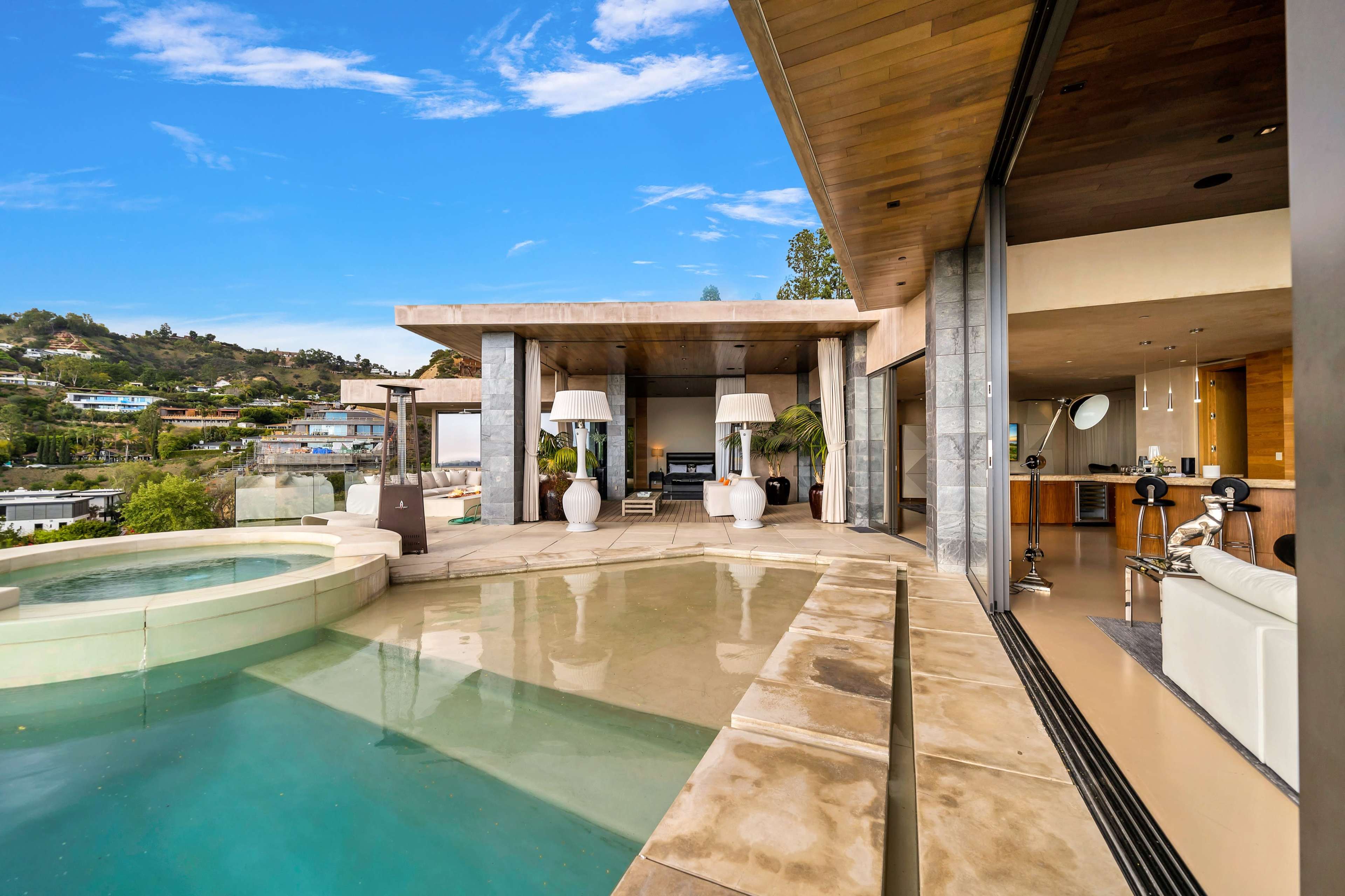 A modern house with large glass walls, a pool area, and stylish furnishings, set against a backdrop of rolling hills.
