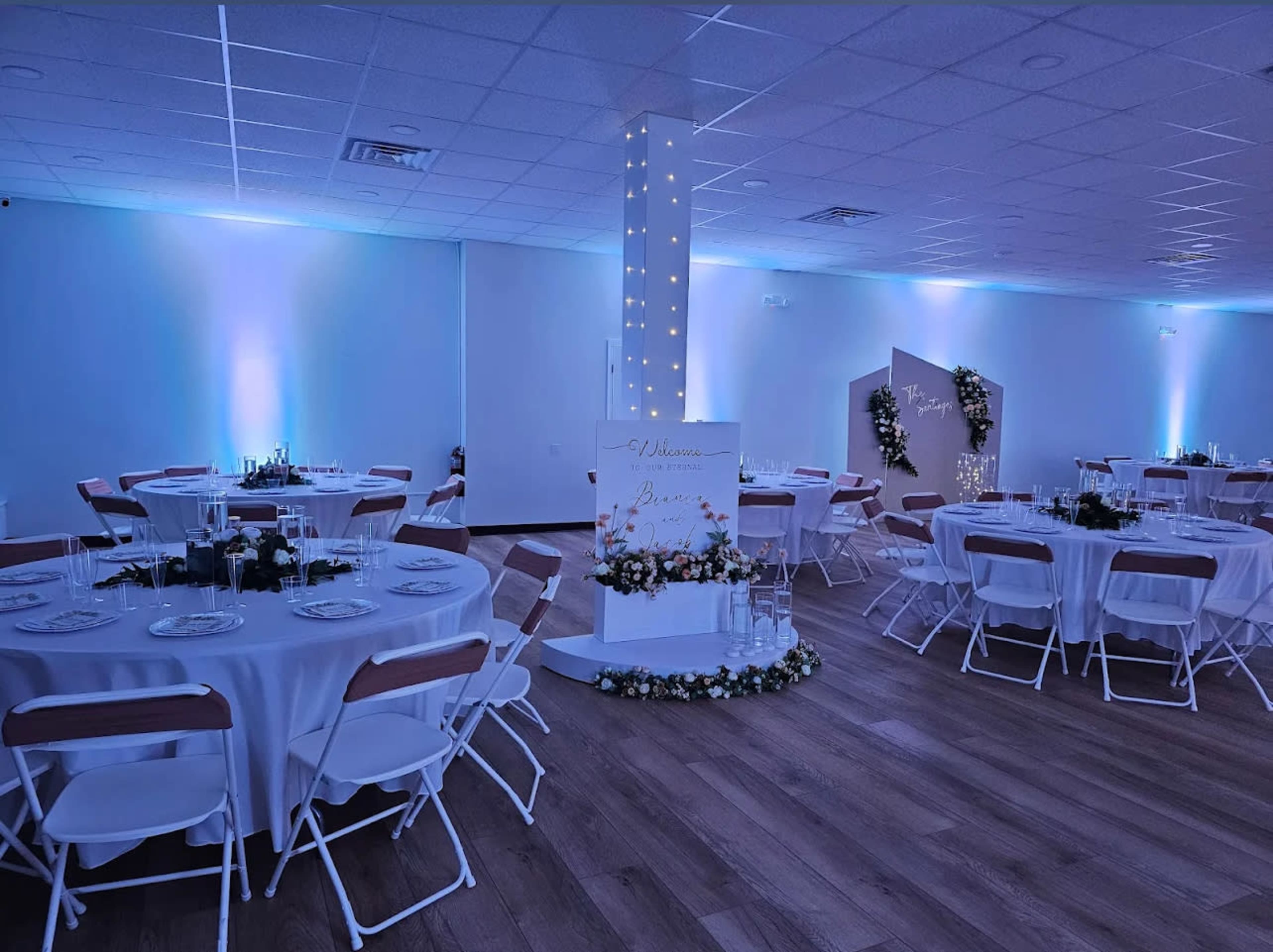 The image shows a well-decorated event space with round tables set for a gathering, illuminated by soft blue lights and adorned with floral arrangements.
