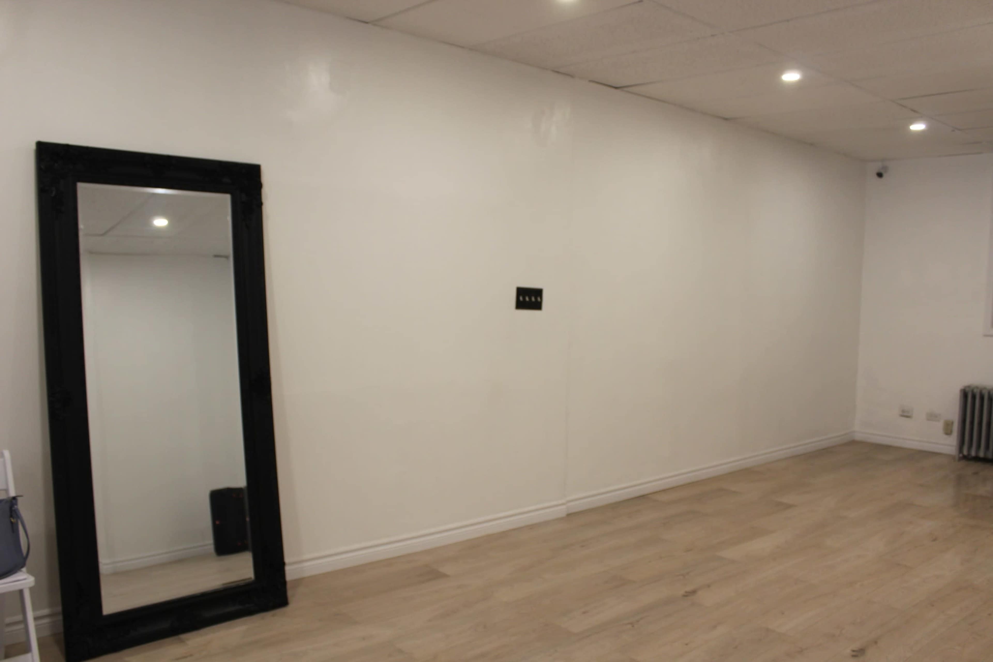 A spacious, empty room with light wooden flooring, white walls, and a large black-framed mirror on one side.