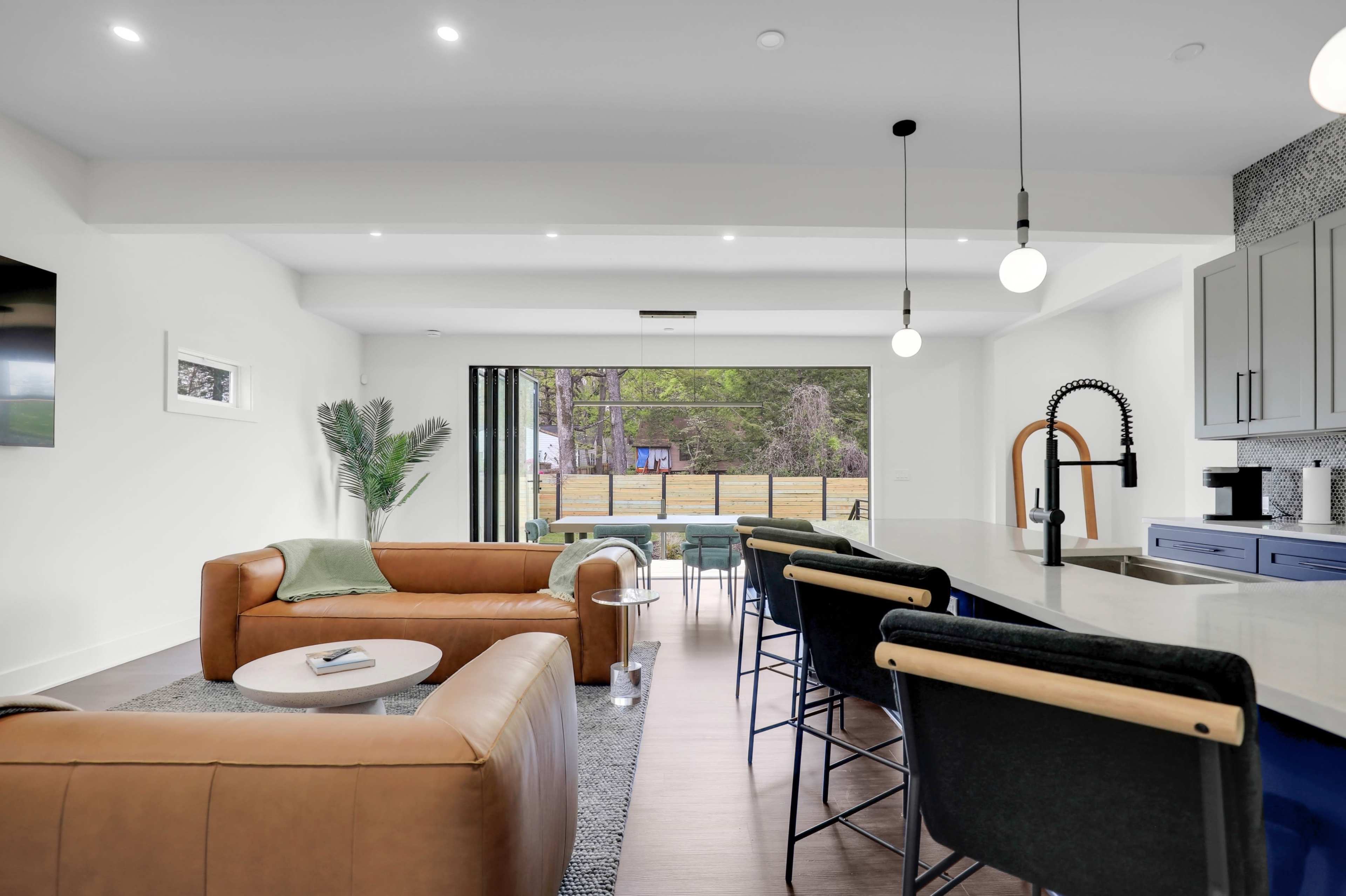 The image shows a modern living space with a brown leather sofa, a light-filled dining area featuring black bar stools, and a large sliding door that opens to an outdoor view.